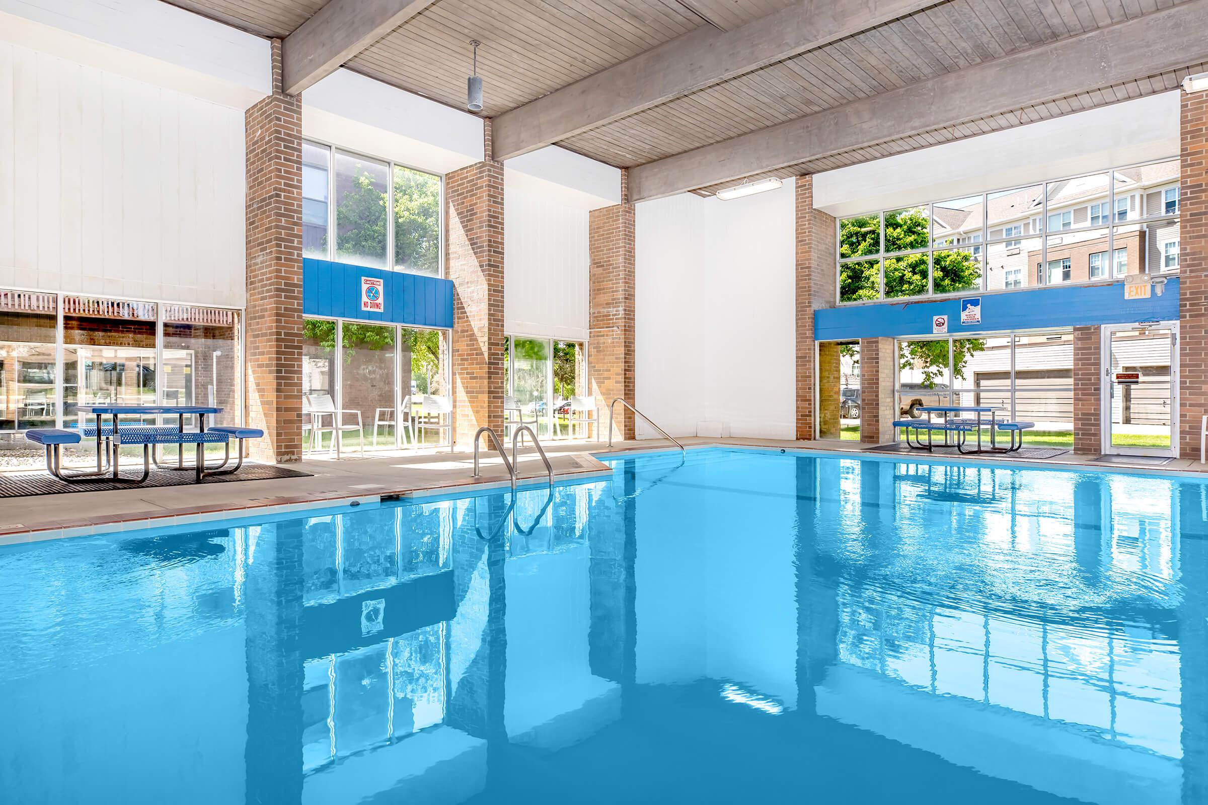 Indoor swimming pool with clear blue water, surrounded by large windows that let in natural light. The pool area features tiled floors and multiple picnic-style tables nearby. The space is bright and inviting, with greenery visible outside the windows.