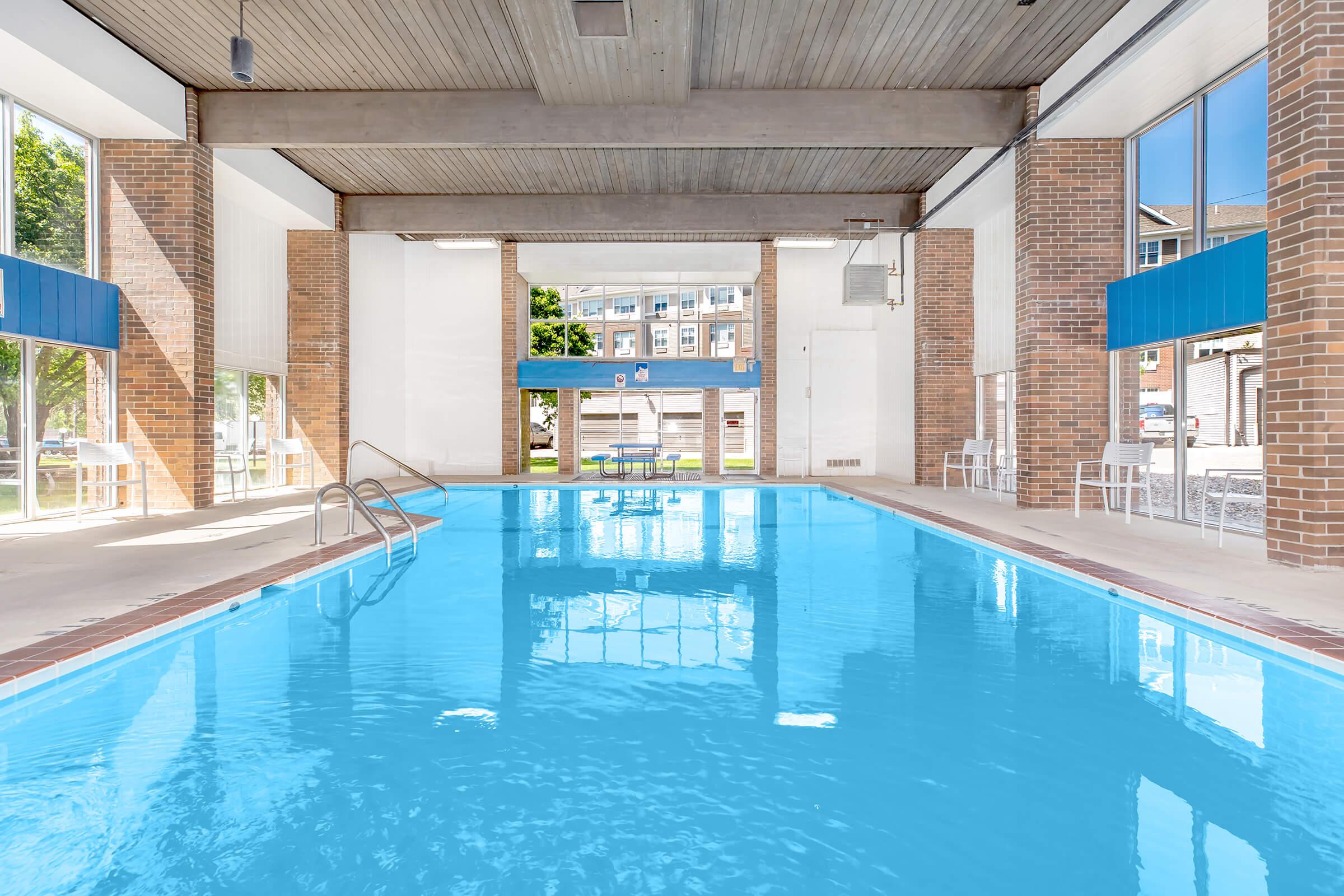 Indoor swimming pool with bright blue water, framed by large windows allowing natural light to flood the space. The room features wooden beams overhead and brick walls, with a few chairs arranged nearby. Reflections on the water create a serene atmosphere.