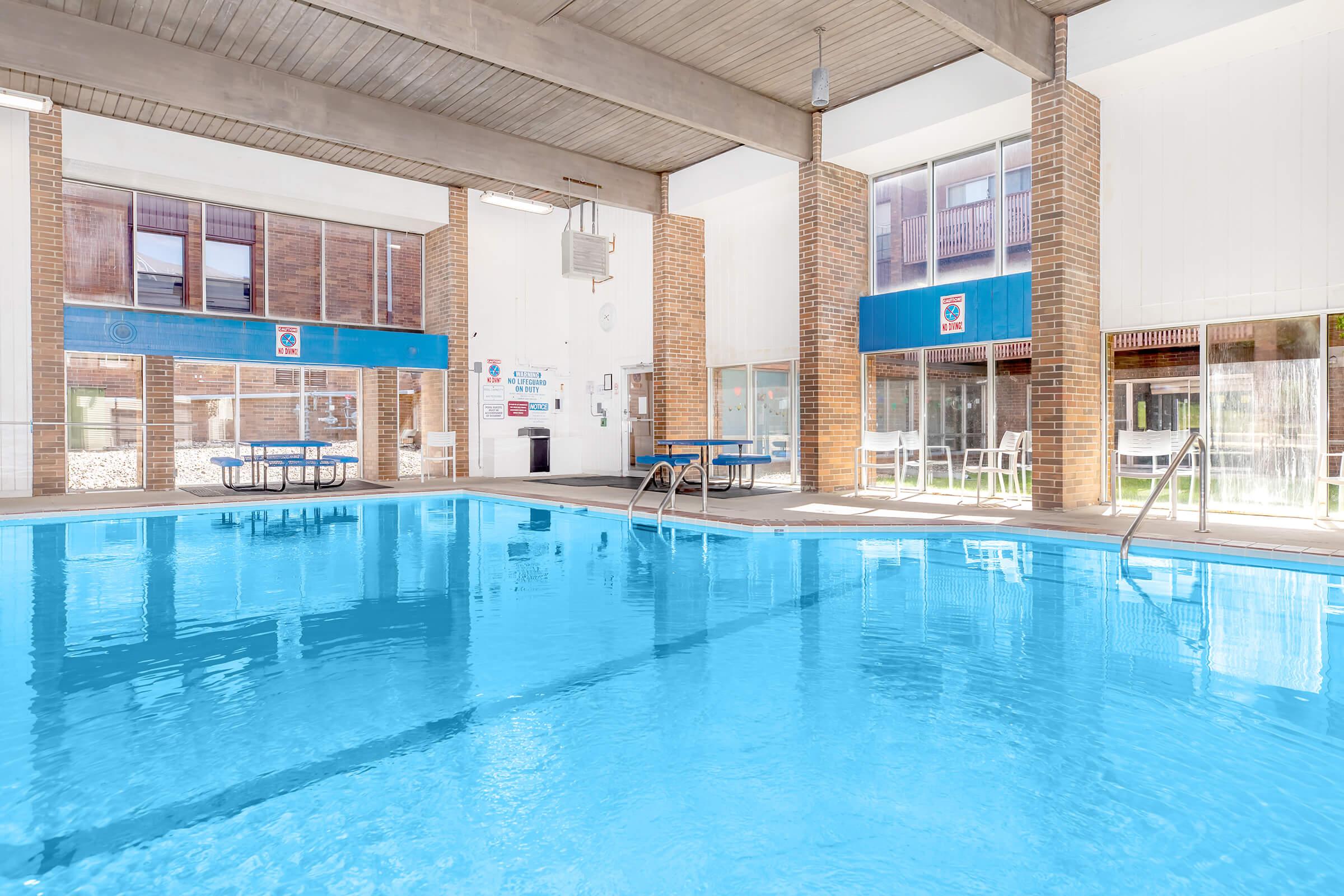 Indoor swimming pool with bright blue water, surrounded by large windows allowing natural light. The area features brick walls and a few tables and chairs. There is signage on the walls and various amenities nearby, creating a clean and inviting atmosphere for relaxation and swimming.