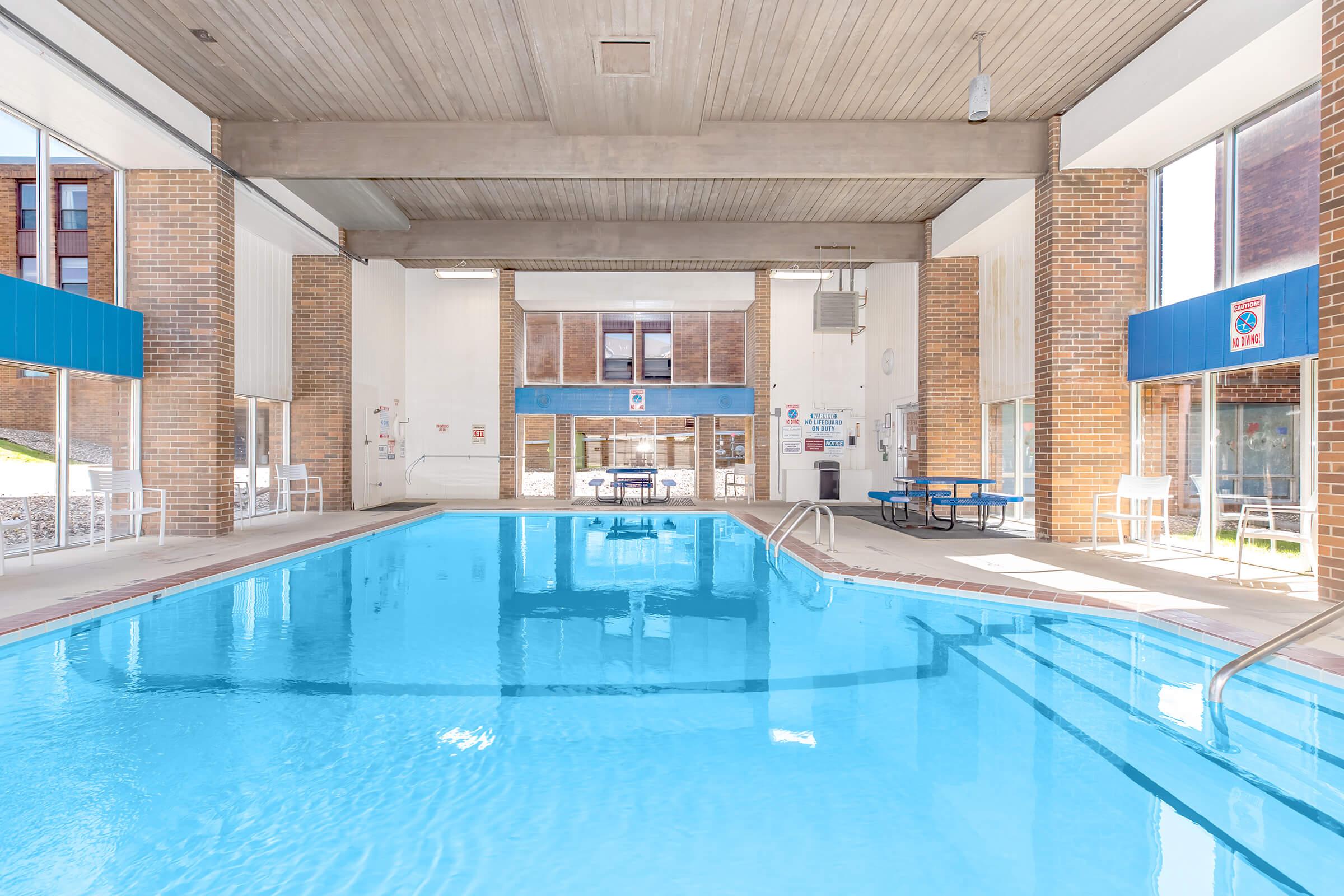 Indoor swimming pool with clear blue water, surrounded by large windows allowing natural light. The walls are of exposed brick and there are tables and chairs nearby. The area appears clean and inviting, perfect for recreational swimming.