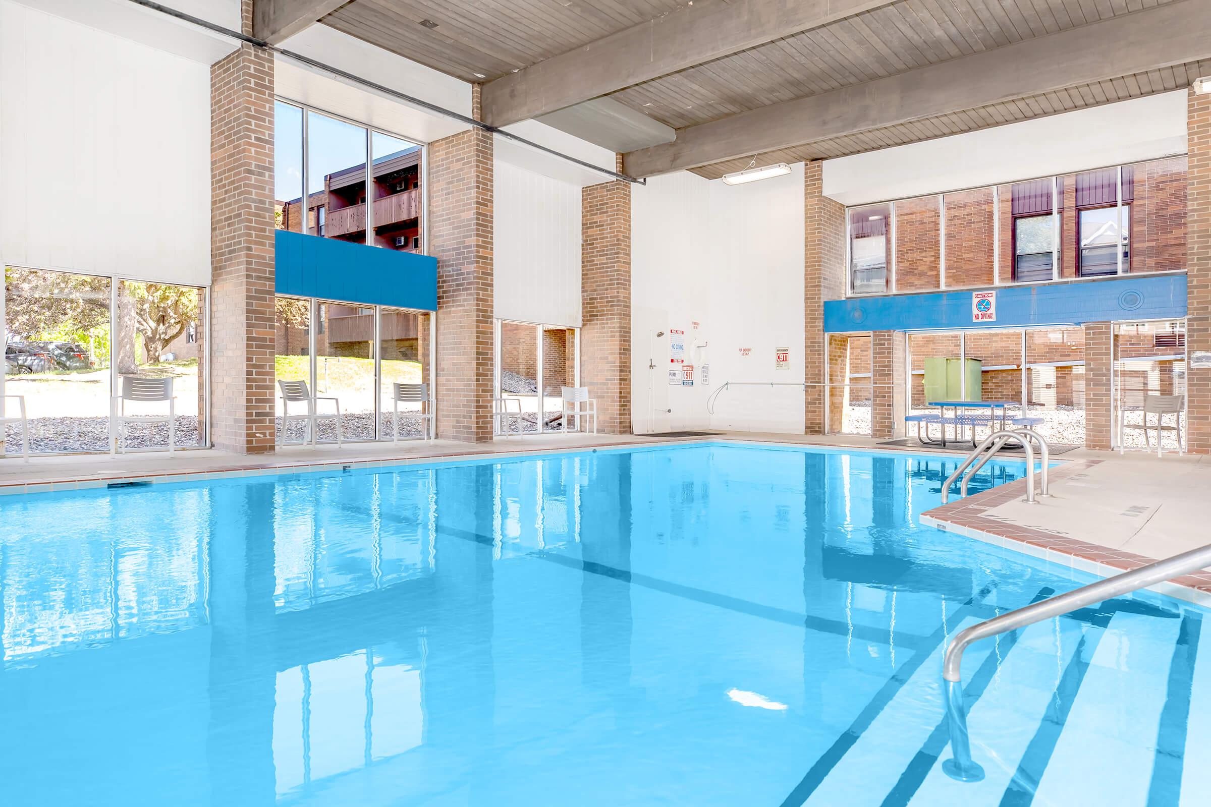 Indoor swimming pool with clear blue water. The area is well-lit, featuring large windows that let in natural light. Brick walls and a clean, modern design give a welcoming atmosphere. Pool steps and handrails are visible, along with chairs positioned around the pool area.