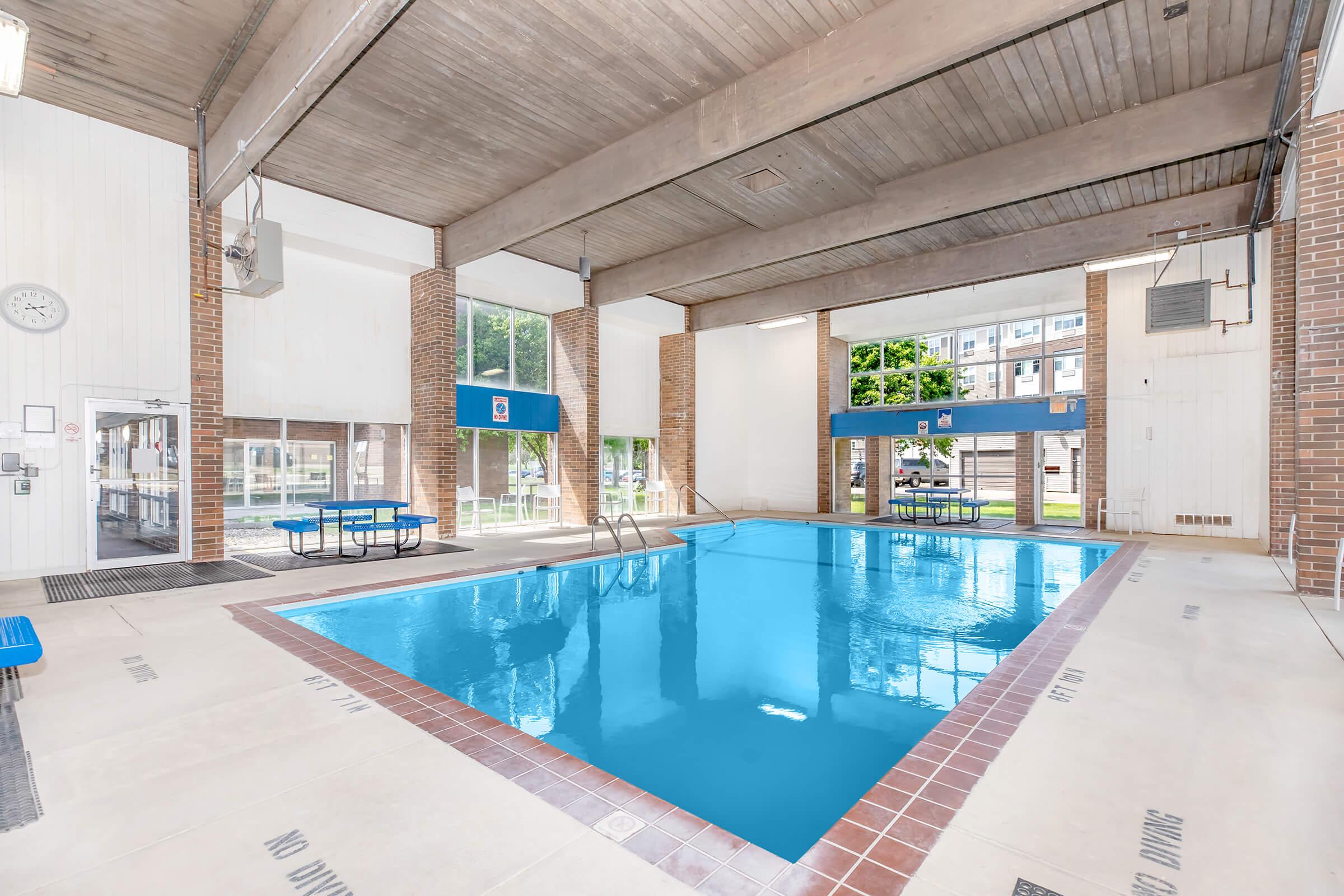 Indoor swimming pool with clear blue water, surrounded by large windows allowing natural light. The pool area features a diving board, tiled surroundings, and seating areas with blue benches. There are pool safety markings on the floor. The atmosphere is bright and inviting.