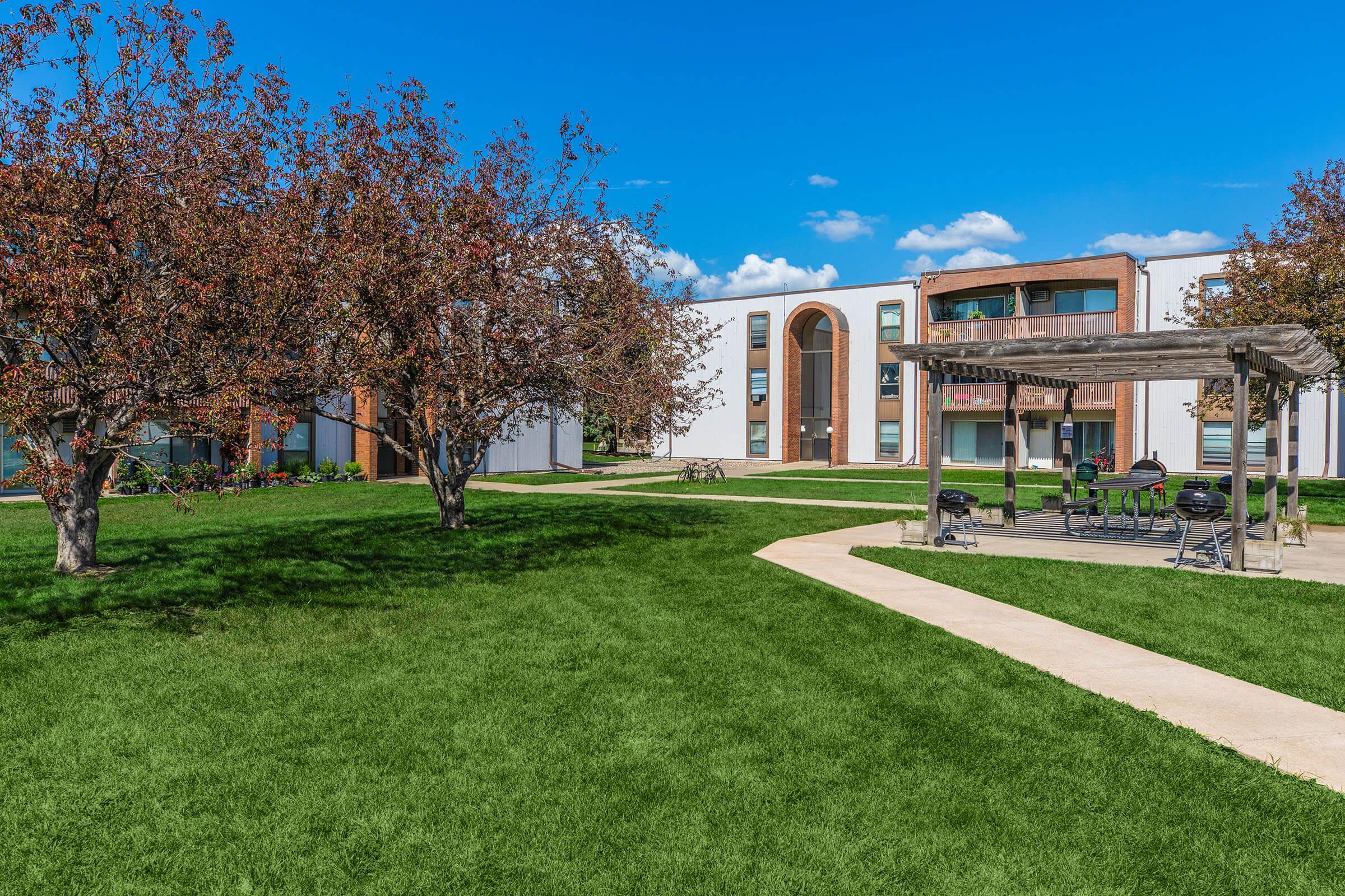 A scenic view of a well-maintained green lawn with blooming trees, a patio area with seating, and a building in the background under a bright blue sky. The scene conveys a peaceful outdoor setting, ideal for relaxation or gatherings.