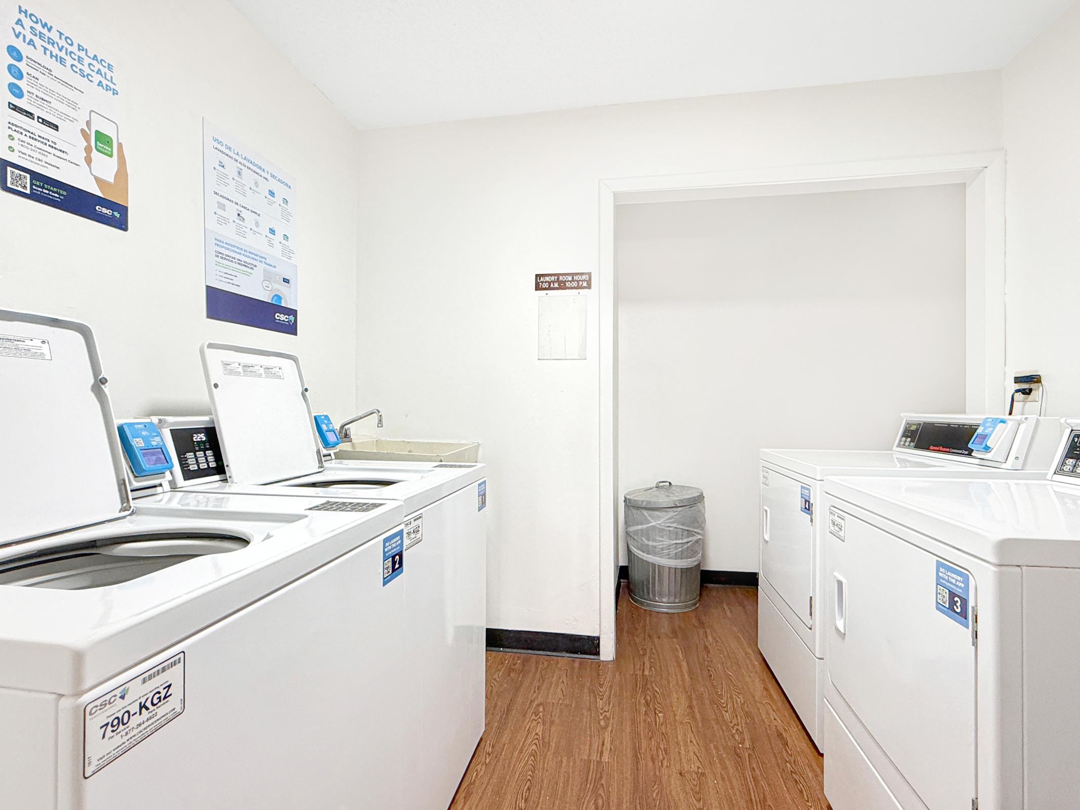 A clean laundry room featuring several white washing machines and dryers. There is a laundry sink along one wall and a trash can in the corner. Wall signage provides instructions for using the machines. The floor is wooden, and the overall space is well-lit and organized.
