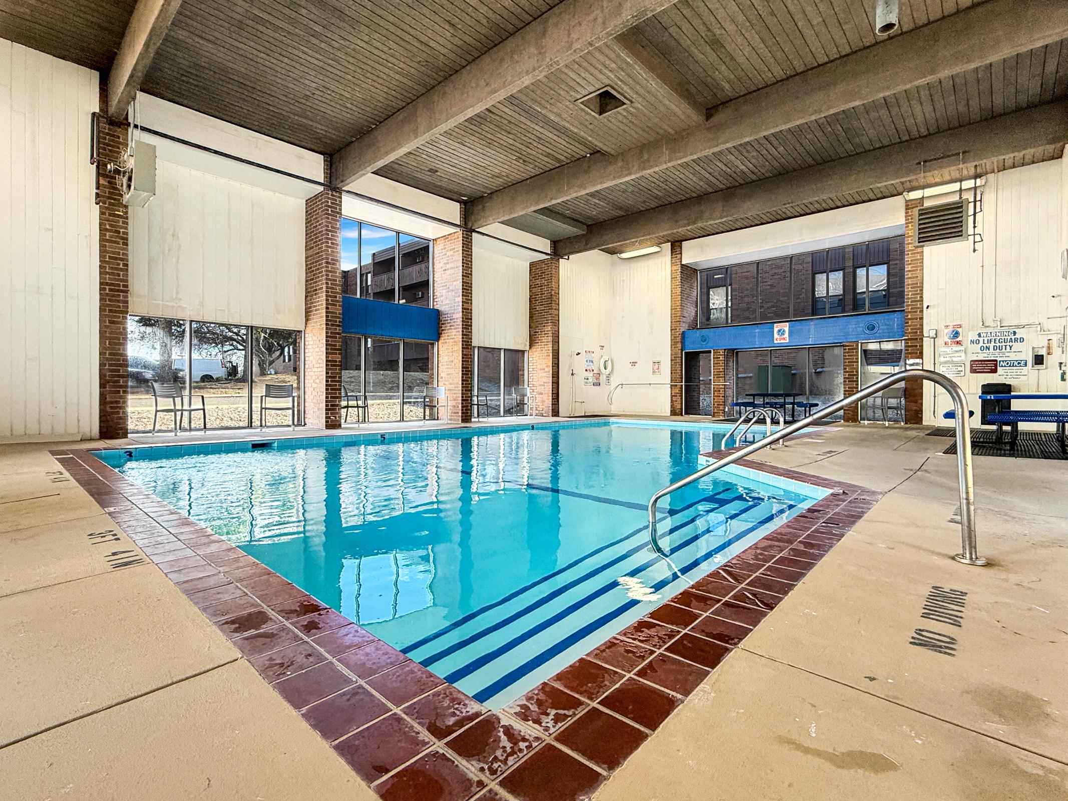 A clean indoor swimming pool with clear blue water, surrounded by white walls and large windows that let in natural light. The pool features a tiled border, a ladder for access, and there are lounge chairs in the background. The atmosphere is bright and inviting, ideal for relaxation or exercise.
