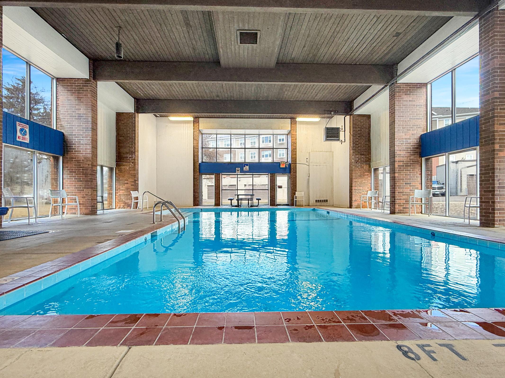 Indoor swimming pool with clear blue water, surrounded by large windows allowing natural light. The pool area features brick walls, white seating, and visible depth markings at the pool's edge. A table is located in the background, adding to the recreational atmosphere.