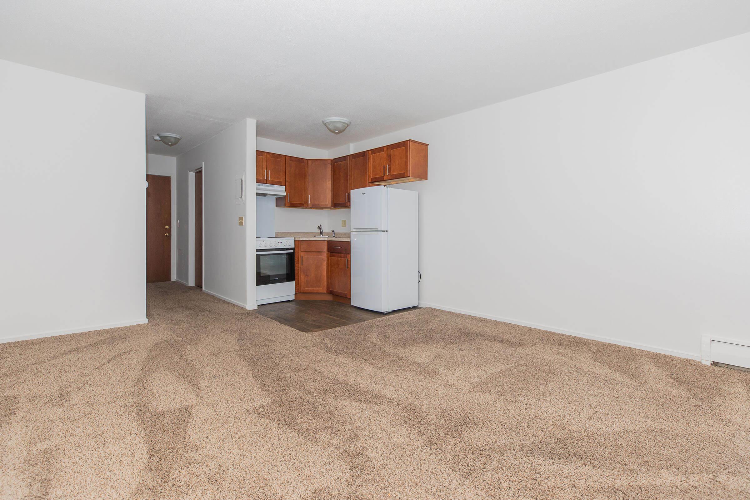 A spacious living area with beige carpet, featuring a small kitchenette in the background equipped with wooden cabinetry, a refrigerator, and an oven. The walls are painted white, and there is a doorway leading to another room on the left. The space is well-lit and empty, ready for furnishing.