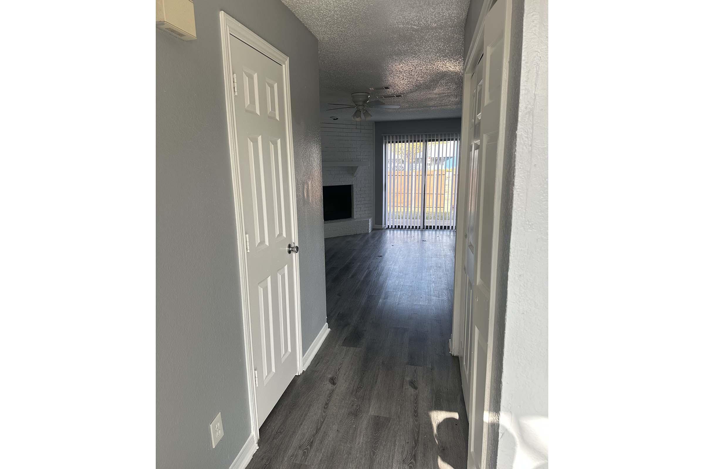 A bright hallway leading into a spacious living area. The walls are painted in light gray, and there are two closed white doors on the left. The living room features large windows with vertical blinds and has a ceiling fan overhead. The flooring is a modern wood-look laminate.