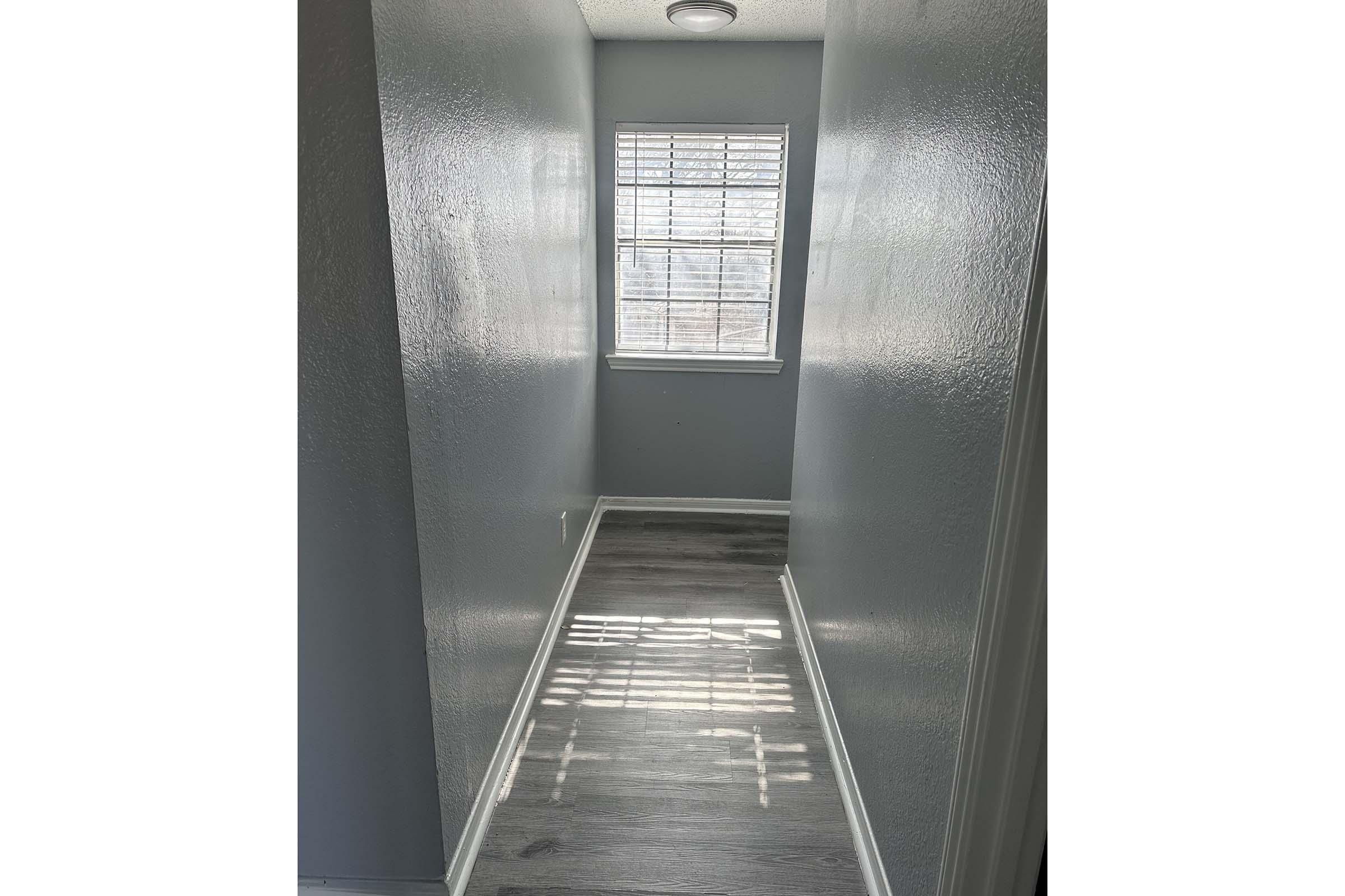 A narrow hallway with light gray walls and a light-colored wooden floor. Sunlight is streaming through a window with white blinds, creating a pattern on the floor. The space is bright and uncluttered, leading to a small room at the end.