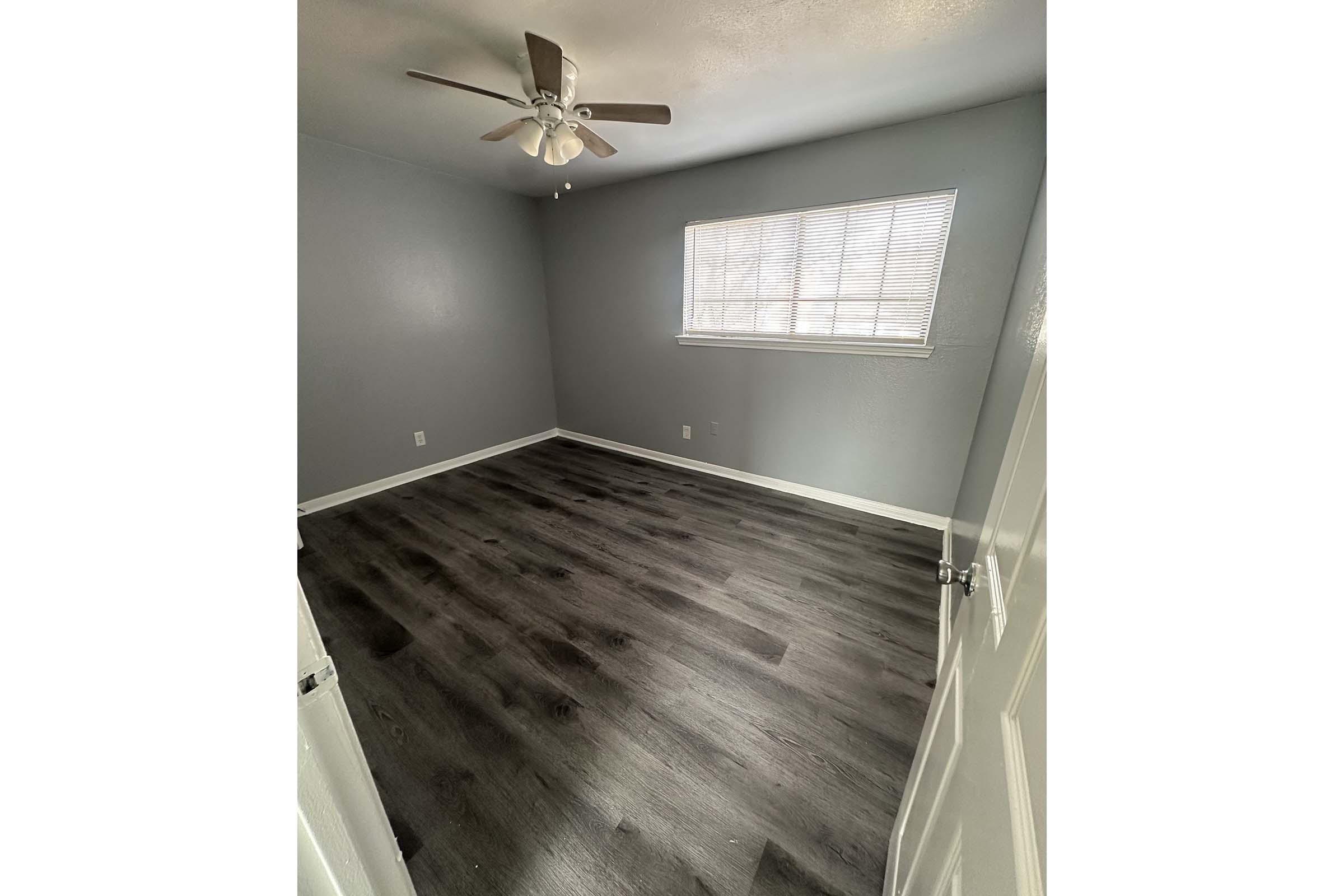 A simple, empty room with gray walls, a ceiling fan, and a window with blinds. The floor is dark-colored laminate, and there are no furnishings or decorations. The entrance door is partially visible on the left side of the image.