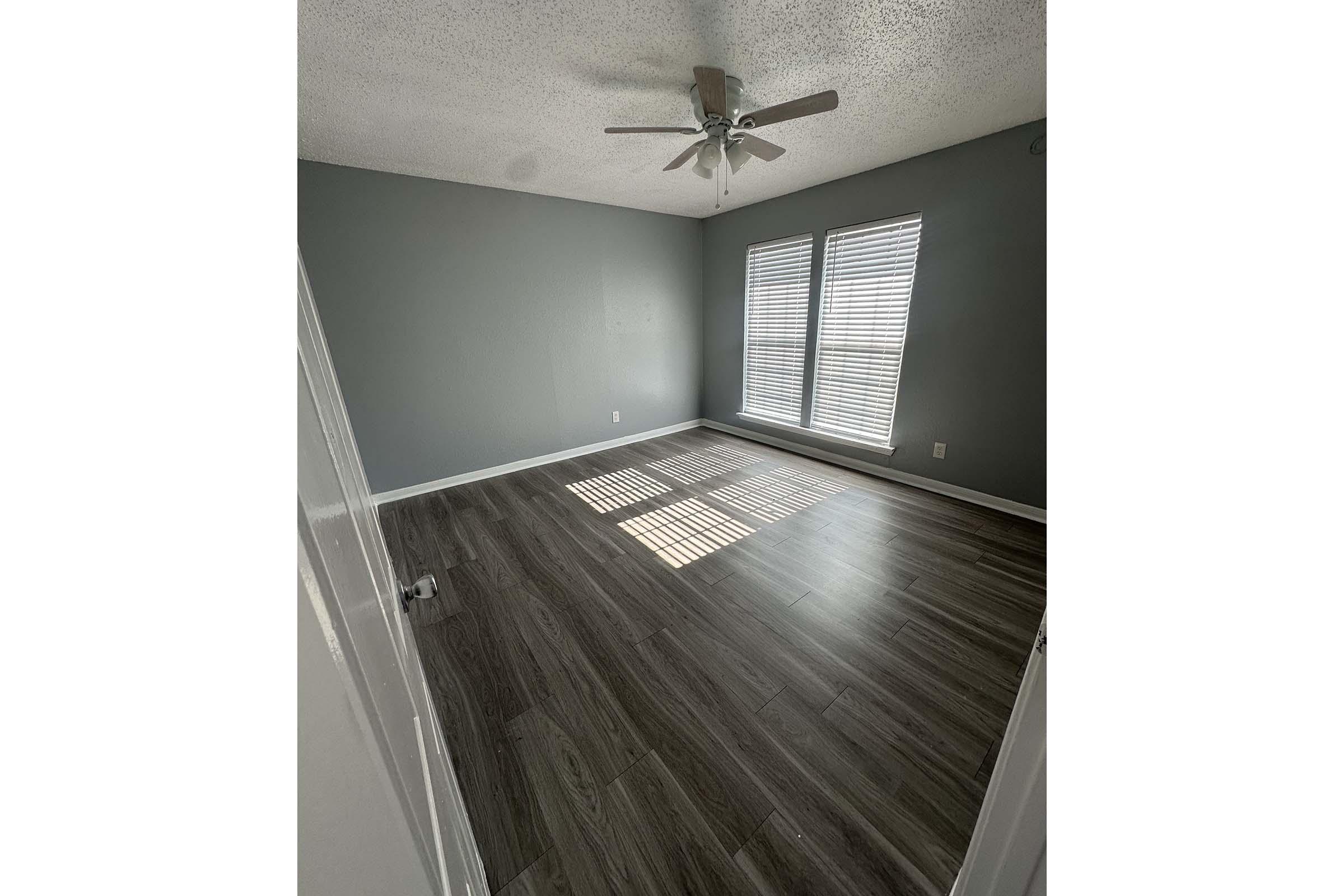 Empty room with light gray walls and dark wood-like flooring. A ceiling fan is visible, and two large windows with white blinds allow natural light to enter. Shadows from the blinds create a striped pattern on the floor. The room is unfurnished and spacious.