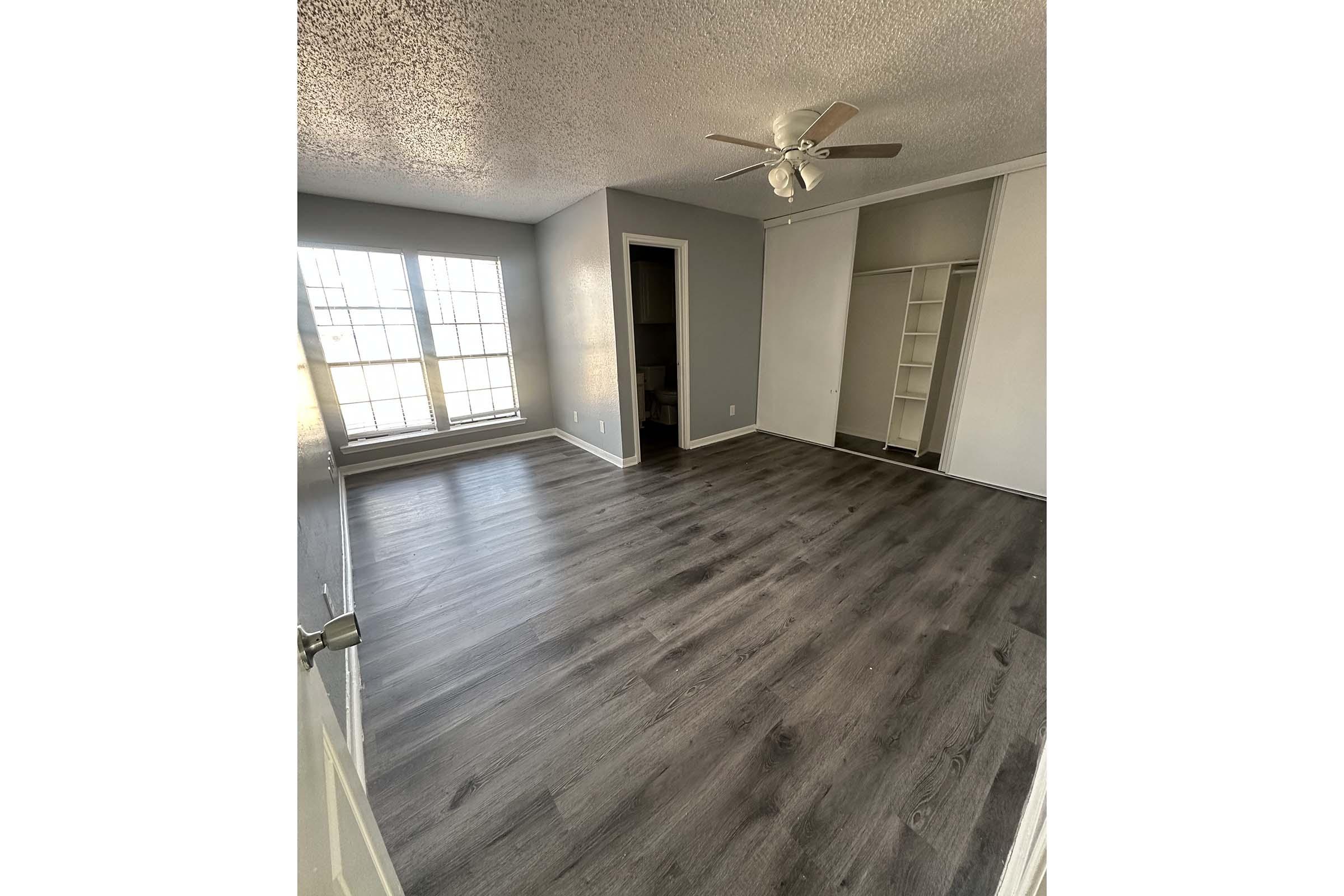 A spacious room with gray walls and large windows letting in natural light. The floor is covered with dark laminate flooring. There is a ceiling fan in the center, and a closet with shelves is visible on one side. The room appears empty and well-lit, creating an inviting atmosphere.