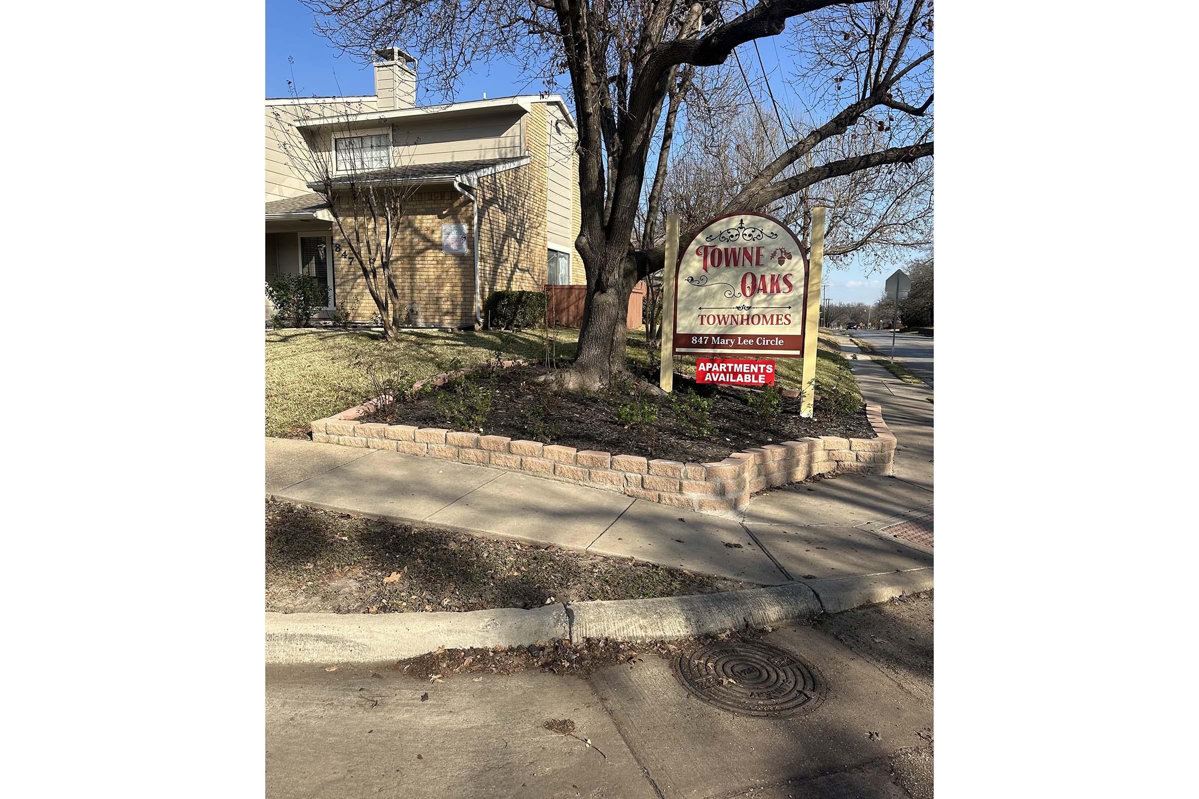 Sign for Towne Oaks Townhomes advertising "Apartments Available," situated in front of a residential building. The sign is near a landscaped area with trees and shrubs, and the setting includes a sidewalk and a partially visible street.