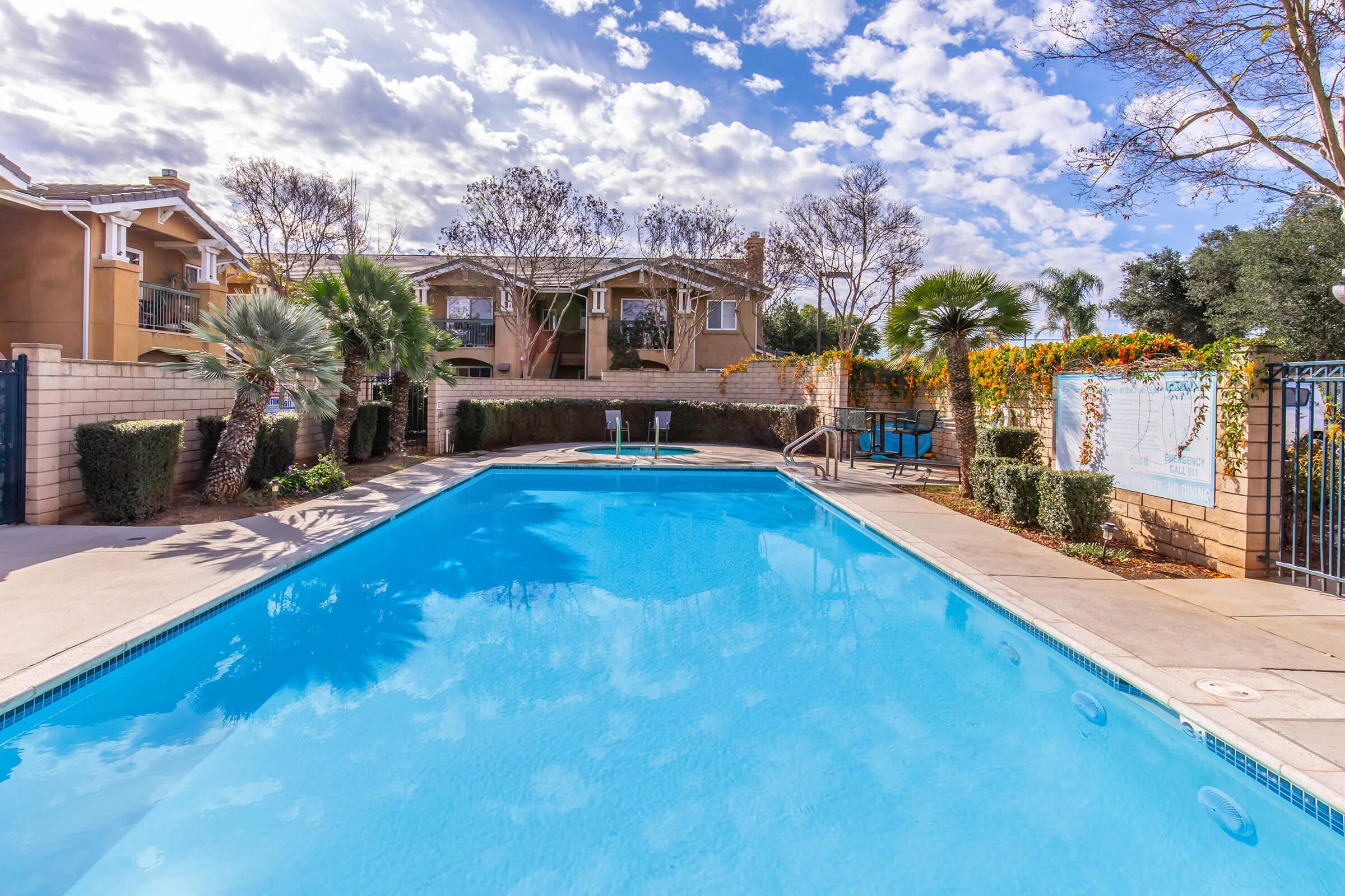 A clear blue swimming pool surrounded by palm trees and apartment buildings, with a fenced area and a cloudy sky overhead. The scene is inviting and sunny, ideal for relaxation.