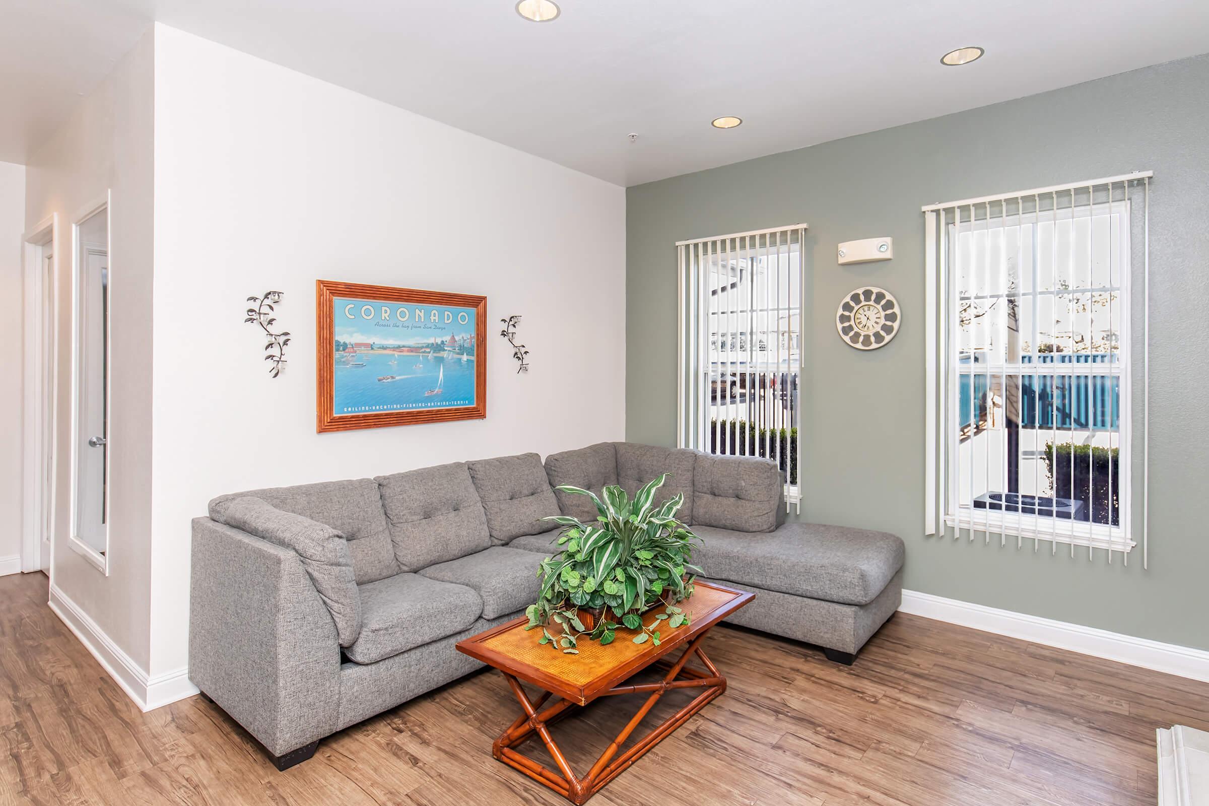 A cozy living room featuring a gray sectional sofa with a green plant on a wooden coffee table. A wall-mounted clock and decorative wall art are present. Natural light flows through two windows adorned with blinds, and there is a subtle coastal theme reflected in the decor.