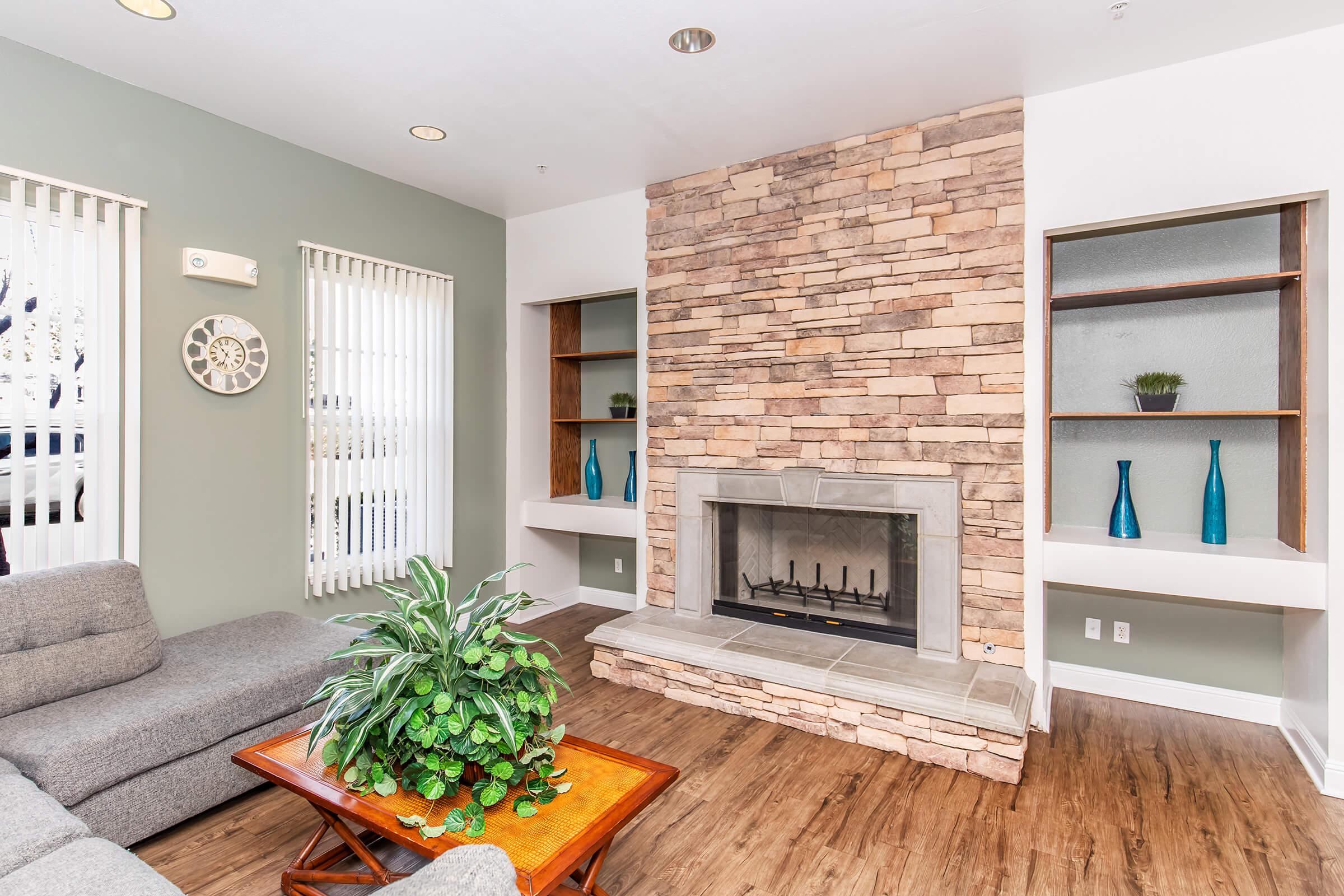 Cozy living room featuring a gray sectional sofa, a wooden coffee table, and a stone fireplace. The walls are painted in soft green, with large windows allowing natural light. Shelves on either side of the fireplace hold decorative vases. A potted plant adds a touch of greenery to the space.