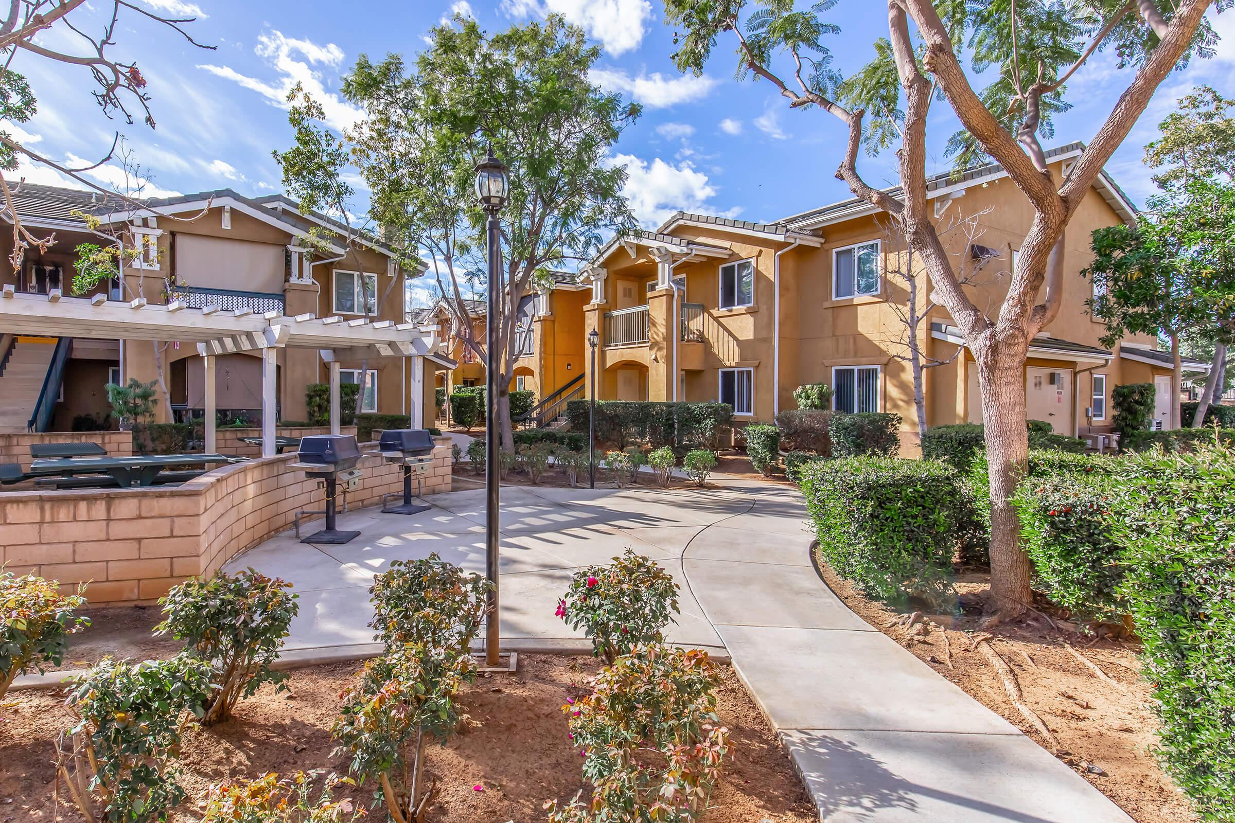 A landscaped area featuring pathways, bushes, and trees, with two residential buildings in the background. The scene includes outdoor seating and barbecues, creating a welcoming communal space. The sky is clear with a few clouds, adding a bright ambiance to the environment.