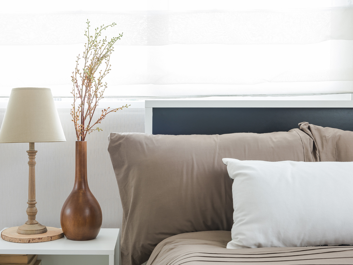 A cozy bedroom setup featuring a neatly made bed with beige and white pillows, a stylish wooden bedside table with a lamp, and a decorative vase with branches. Soft, light curtains allow natural light to filter in, creating a warm and inviting atmosphere.
