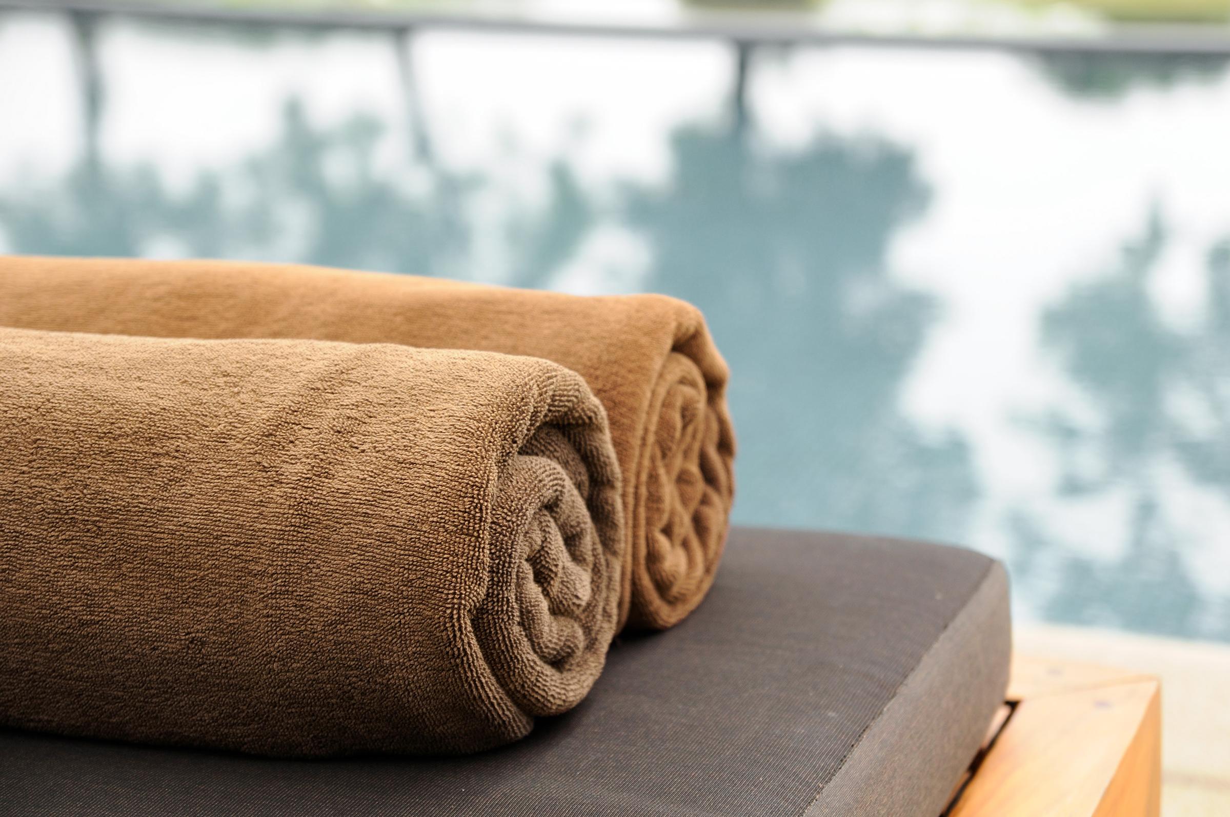 Two neatly rolled brown towels placed on a dark cushion, beside a calm swimming pool with reflections of trees in the water. The scene conveys a tranquil, relaxing atmosphere ideal for leisure and self-care.