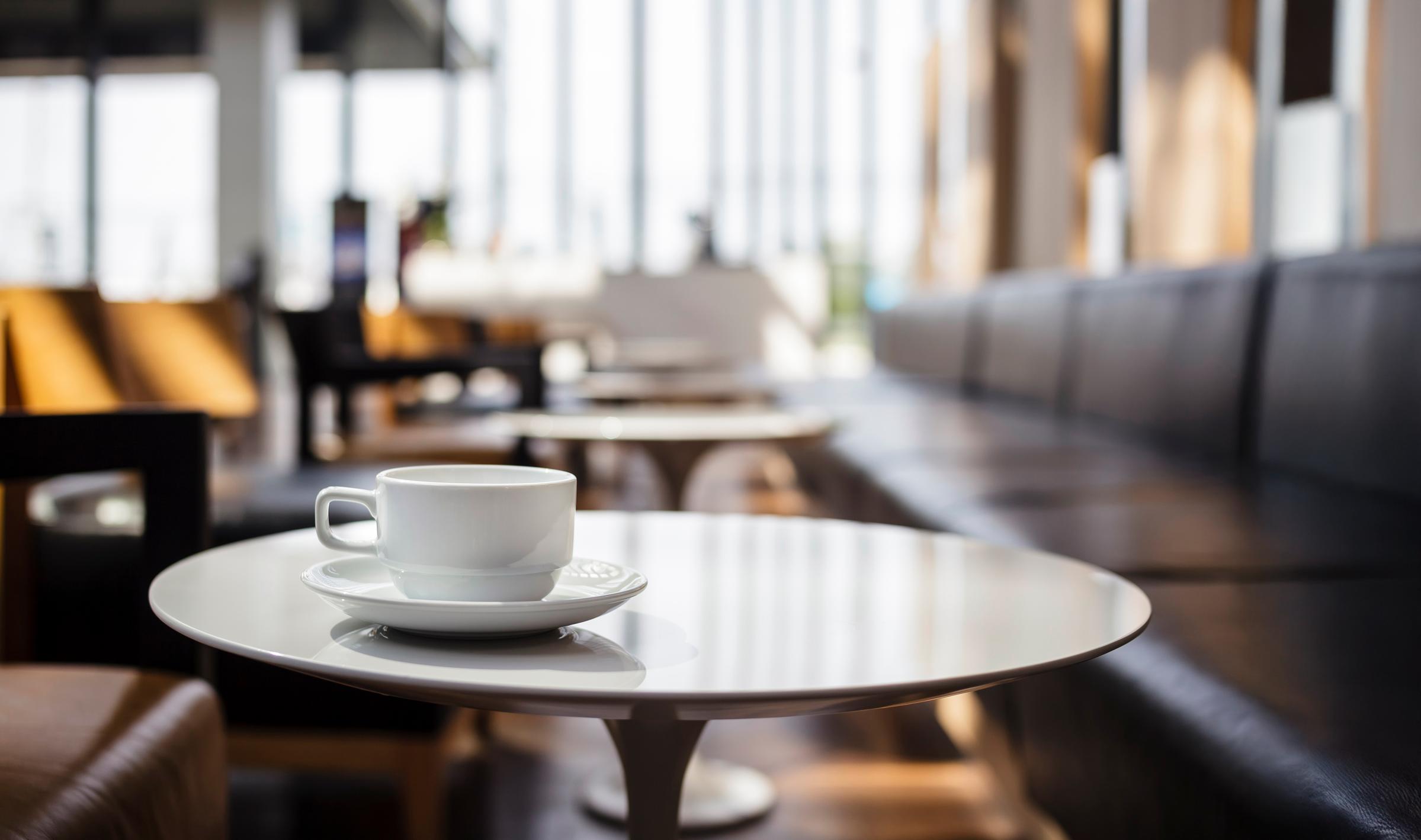 A white coffee cup on a saucer sits on a small table in a modern, brightly lit café. The background features a row of empty seats and large windows allowing natural light to flood the space. The atmosphere is calm and inviting, ideal for enjoying a quiet moment with a beverage.