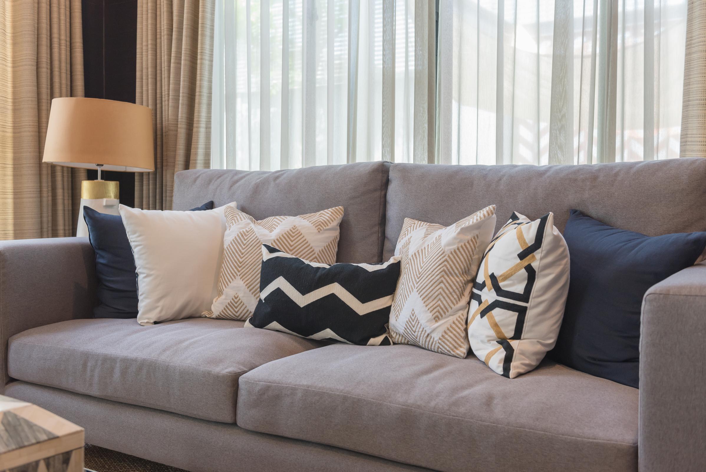 A cozy gray sofa adorned with a variety of decorative pillows in different patterns and colors, including black, white, and gold accents. Natural light filters through sheer curtains in the background, creating a warm and inviting atmosphere. A stylish lamp sits beside the sofa.
