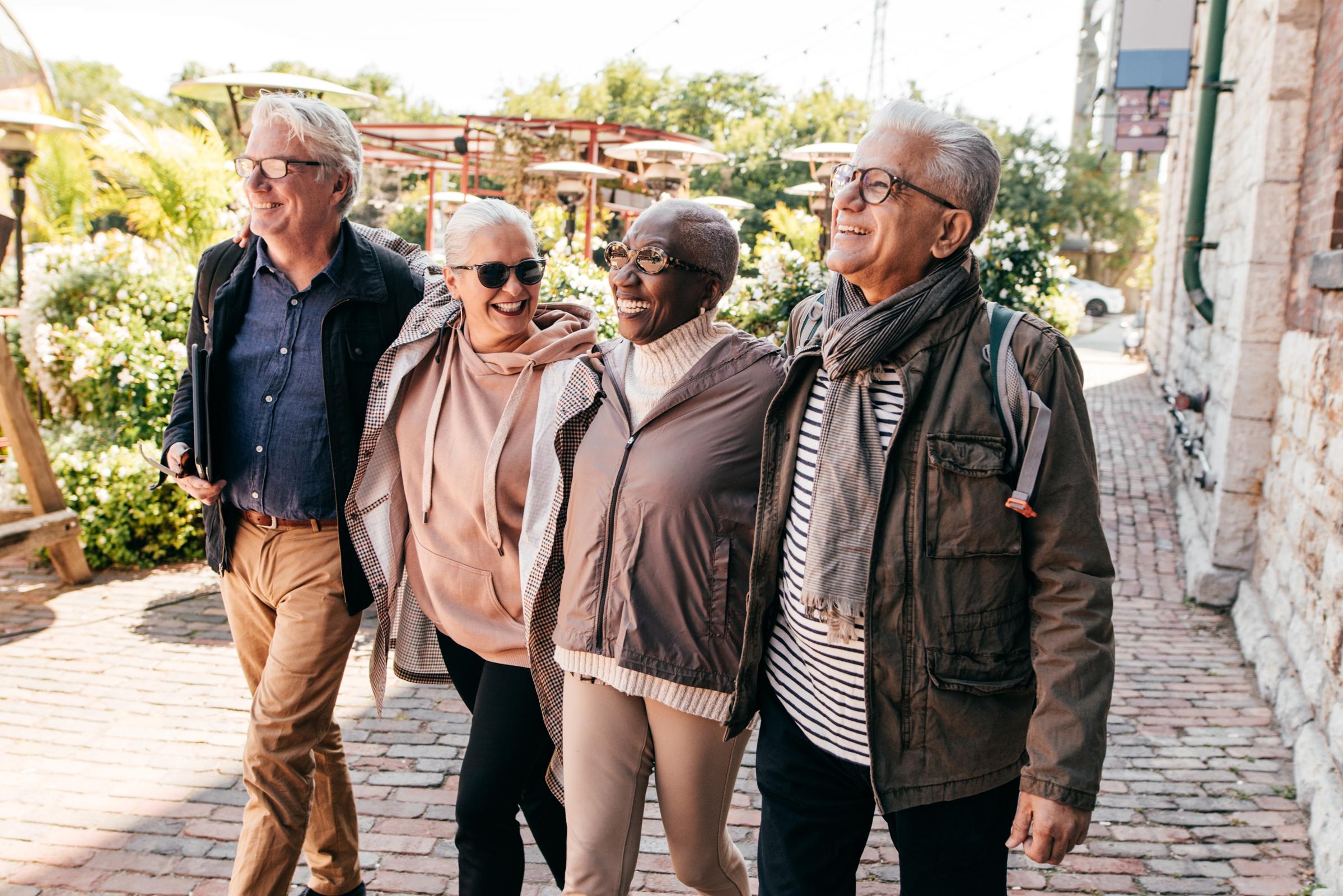 A diverse group of four older adults walking together outdoors, smiling and enjoying each other's company. They are dressed in casual attire and appear to be in a vibrant, lively setting with greenery and outdoor seating in the background.