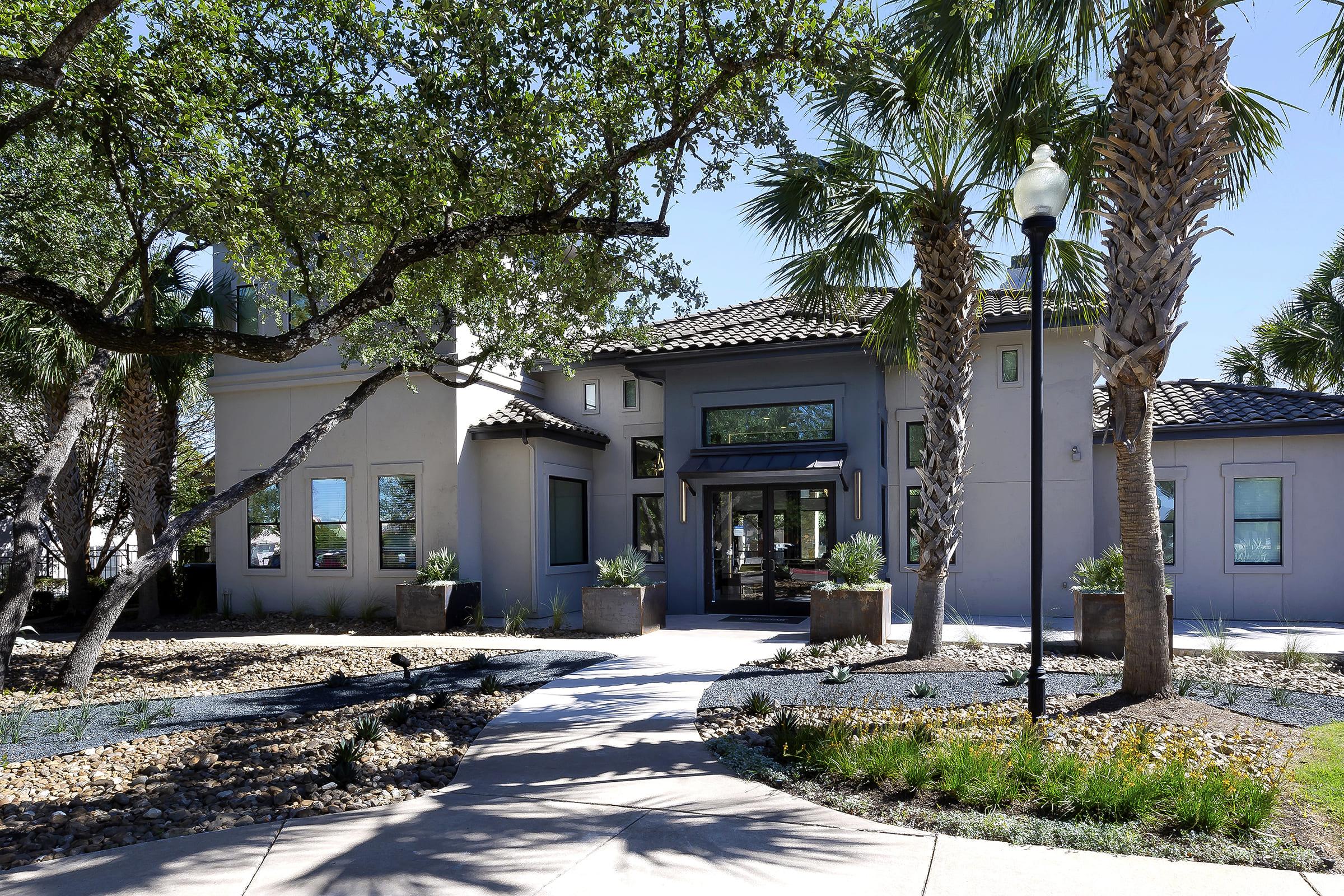 A modern, two-story building surrounded by palm trees and lush greenery. The entrance features large windows and a glass door, with a pathway leading to it. Decorative landscaping includes rocks and small plants, creating a welcoming outdoor space.