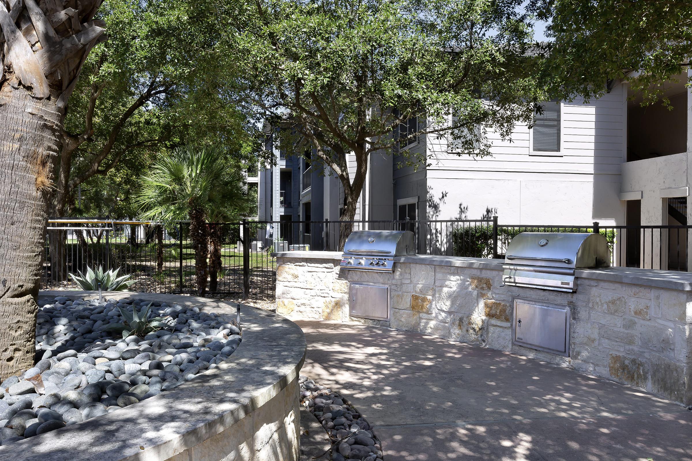 Outdoor grill area with two stainless steel BBQ grills set into a stone structure. Surrounding the grills are smooth gray stones and landscaping featuring palm trees and other greenery. In the background, there are modern apartment buildings partially visible behind a black fence. A bright, sunny day enhances the scene.