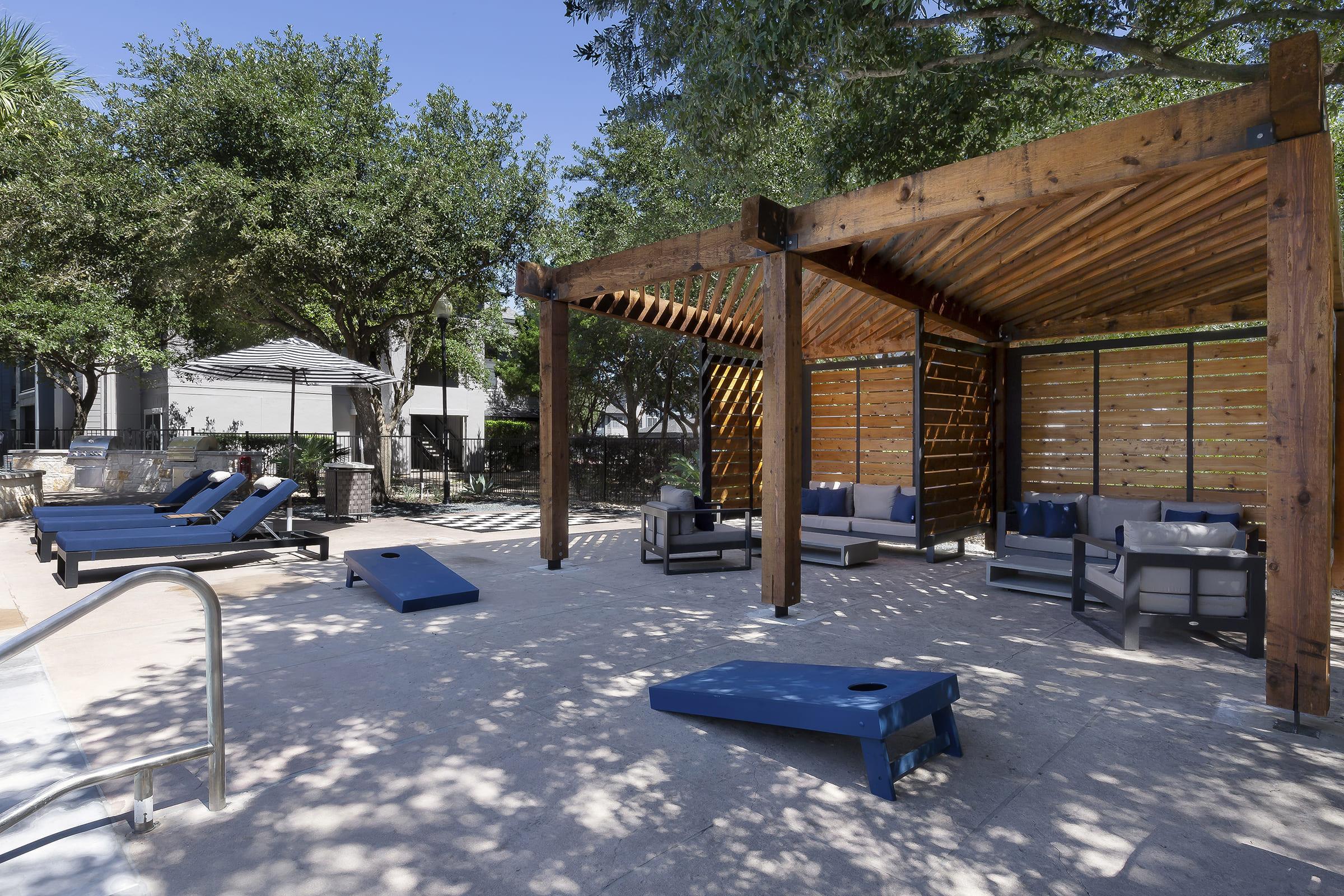 A sunny outdoor space featuring wooden structures, lounge chairs, and seating areas. There’s a clean, paved area with cornhole boards and lush trees providing shade, creating a relaxing atmosphere for leisure and social gatherings.