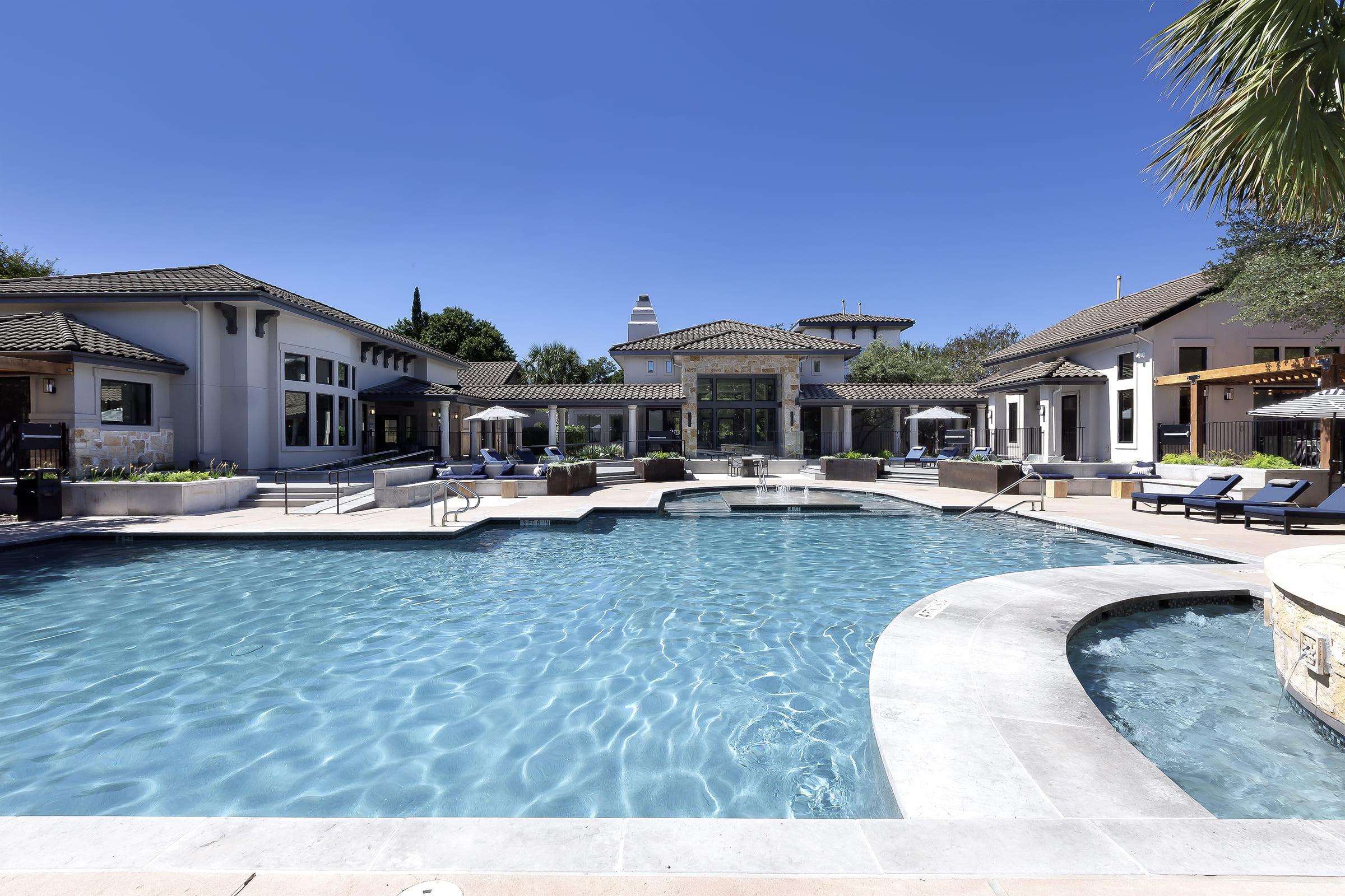 A spacious outdoor pool area surrounded by lounge chairs and umbrellas, with a modern clubhouse in the background. The scene features clear blue skies and well-maintained landscaping, creating a relaxing atmosphere ideal for leisure and social gatherings.