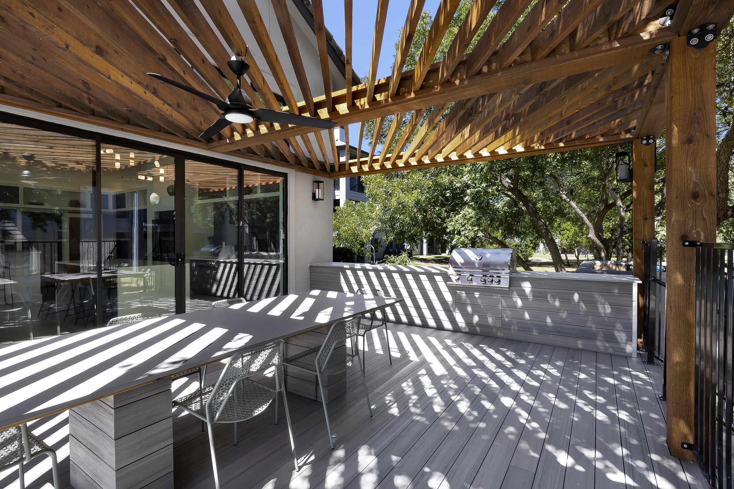 A modern outdoor patio area with a wooden pergola casting striped shadows. The space features a large table with metal chairs, surrounded by greenery. Windows provide a view of the interior, and there’s a grill on the side, creating an inviting atmosphere for dining or relaxation.