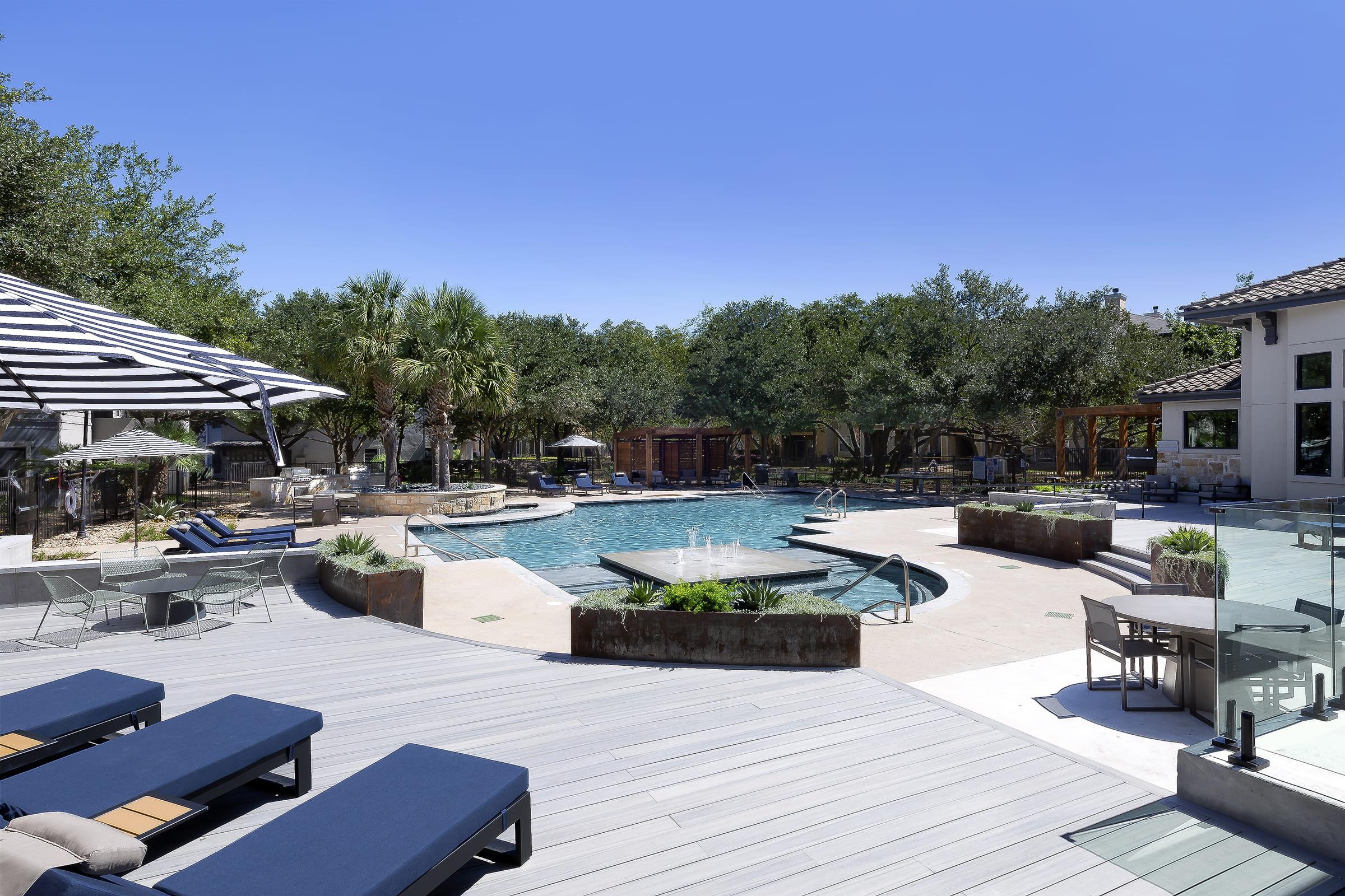 A luxurious outdoor pool area featuring a large blue pool surrounded by sun loungers and tropical landscaping. Umbrellas provide shade, and there are tables and chairs for relaxation. The space is bright and inviting, with clear blue skies and a well-maintained environment ideal for leisure activities.