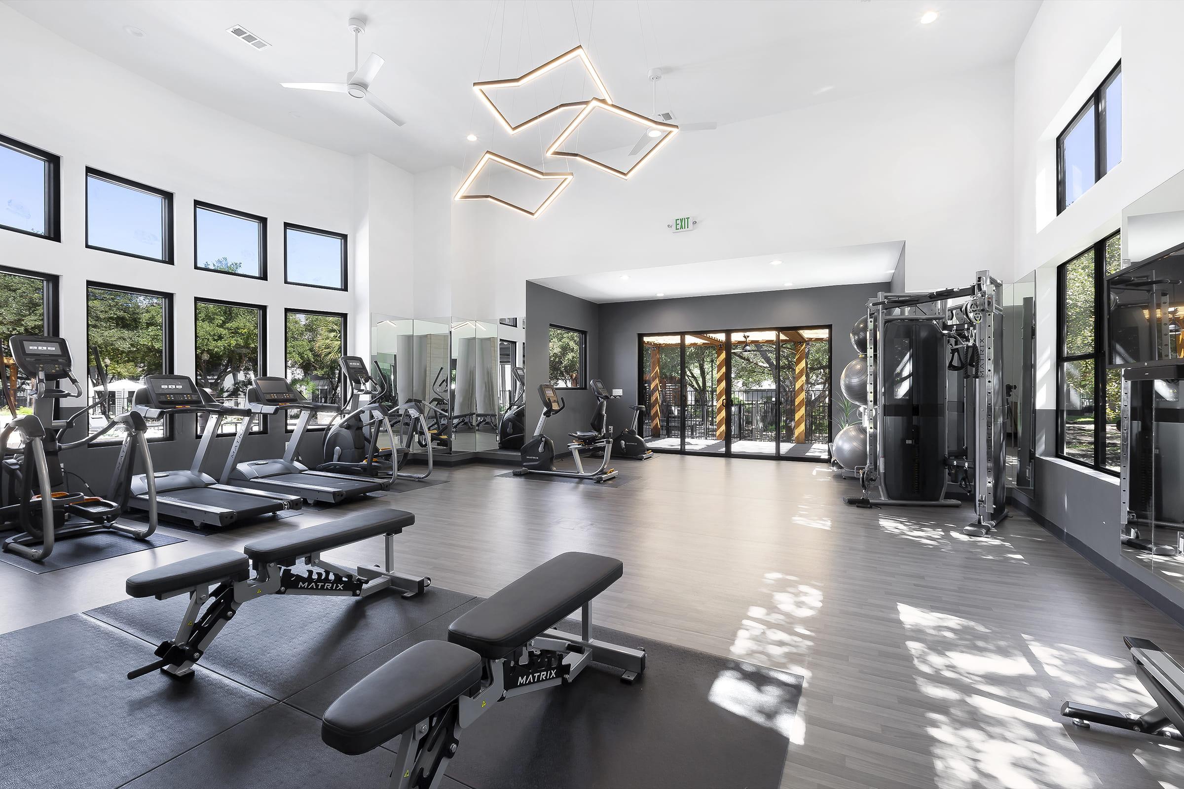 A modern gym interior featuring rows of treadmills and exercise bikes, with large windows allowing natural light. There are weight benches and a cable machine in the background, surrounded by a sleek, minimalist design. The space has a contemporary lighting fixture and views of greenery outside.