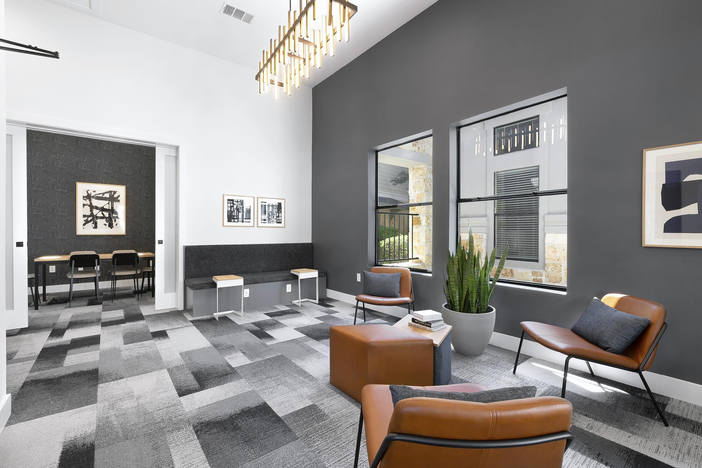 A modern lounge area featuring two brown leather chairs, a large potted plant, and a gray accent wall. The space includes a patterned carpet and large windows allowing natural light. Decorative artwork lines the walls, and a small table with books is positioned nearby. A dining area is visible through an open doorway.