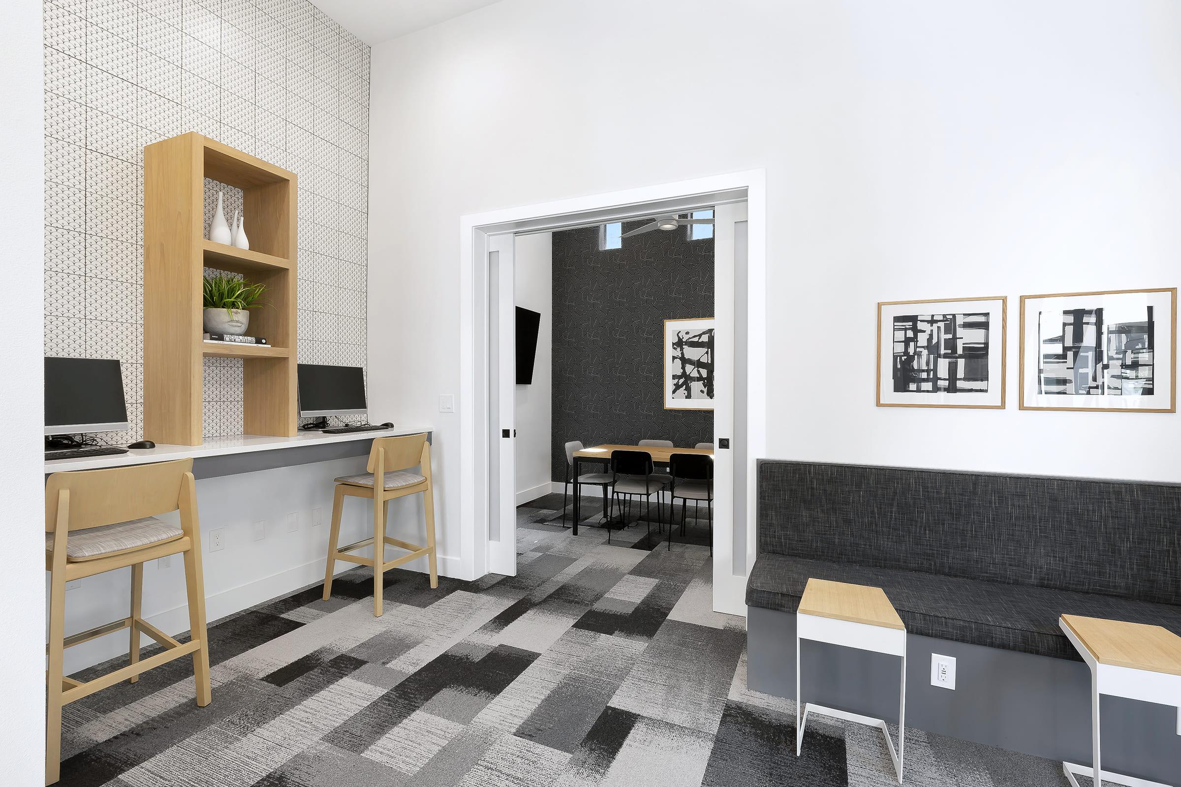 Modern workspace featuring two computers on a light wooden desk, surrounded by patterned gray walls. A small meeting area with a dark gray wall and abstract art is visible. There’s a gray seating area with two light wood side tables. The overall decor is minimalist and contemporary.