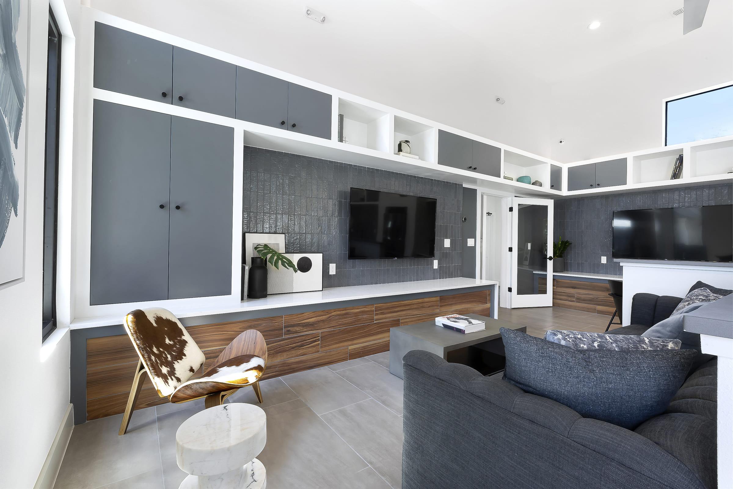 Modern living room with a stylish design featuring gray and wooden cabinetry, two flat-screen TVs mounted on the wall, and a comfortable gray sofa. A unique patterned chair and a small round table are positioned nearby, creating an inviting and contemporary atmosphere. Large windows allow natural light to fill the space.