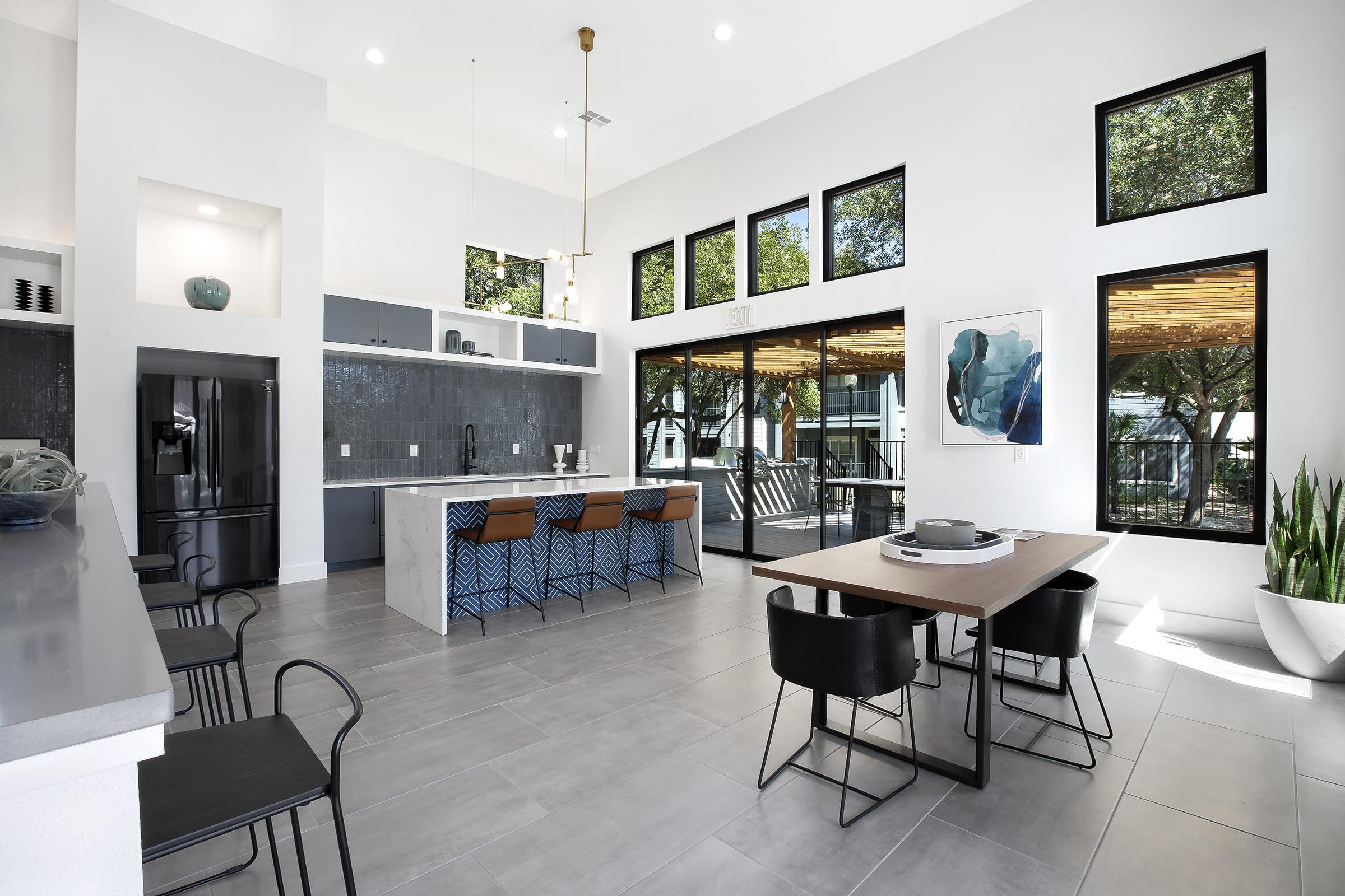 Modern kitchen and dining area with large windows, featuring a central island with bar stools, a dining table with black chairs, and a sleek design. The space is bright and airy, with a minimalist aesthetic and greenery visible outside through the glass doors.