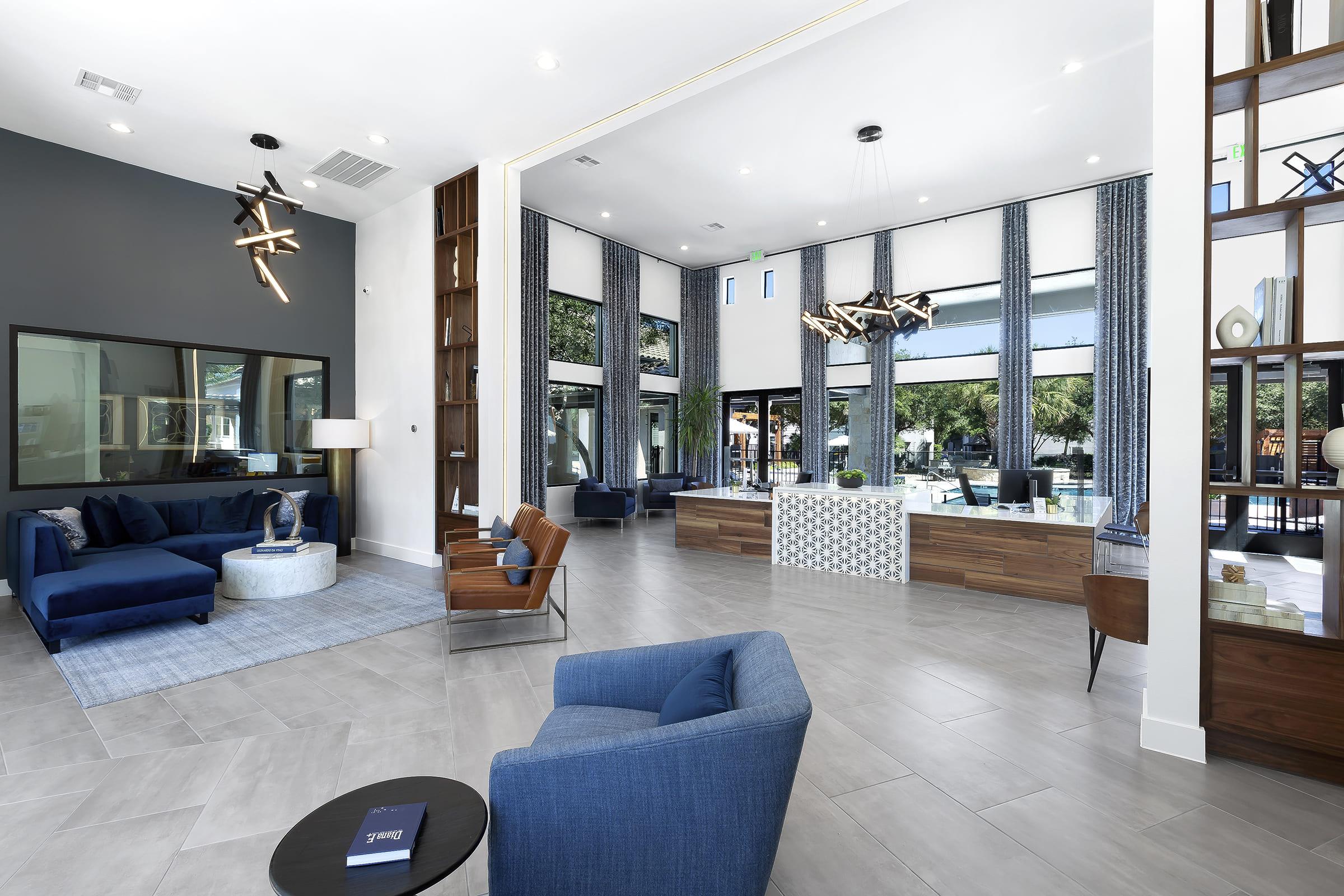 A modern lobby interior featuring stylish seating arrangements with blue sofas and wooden chairs. Large windows allow natural light, and decorative light fixtures add a contemporary touch. A reception area with a wooden desk is positioned prominently. Neutral-colored walls and flooring create an inviting atmosphere.