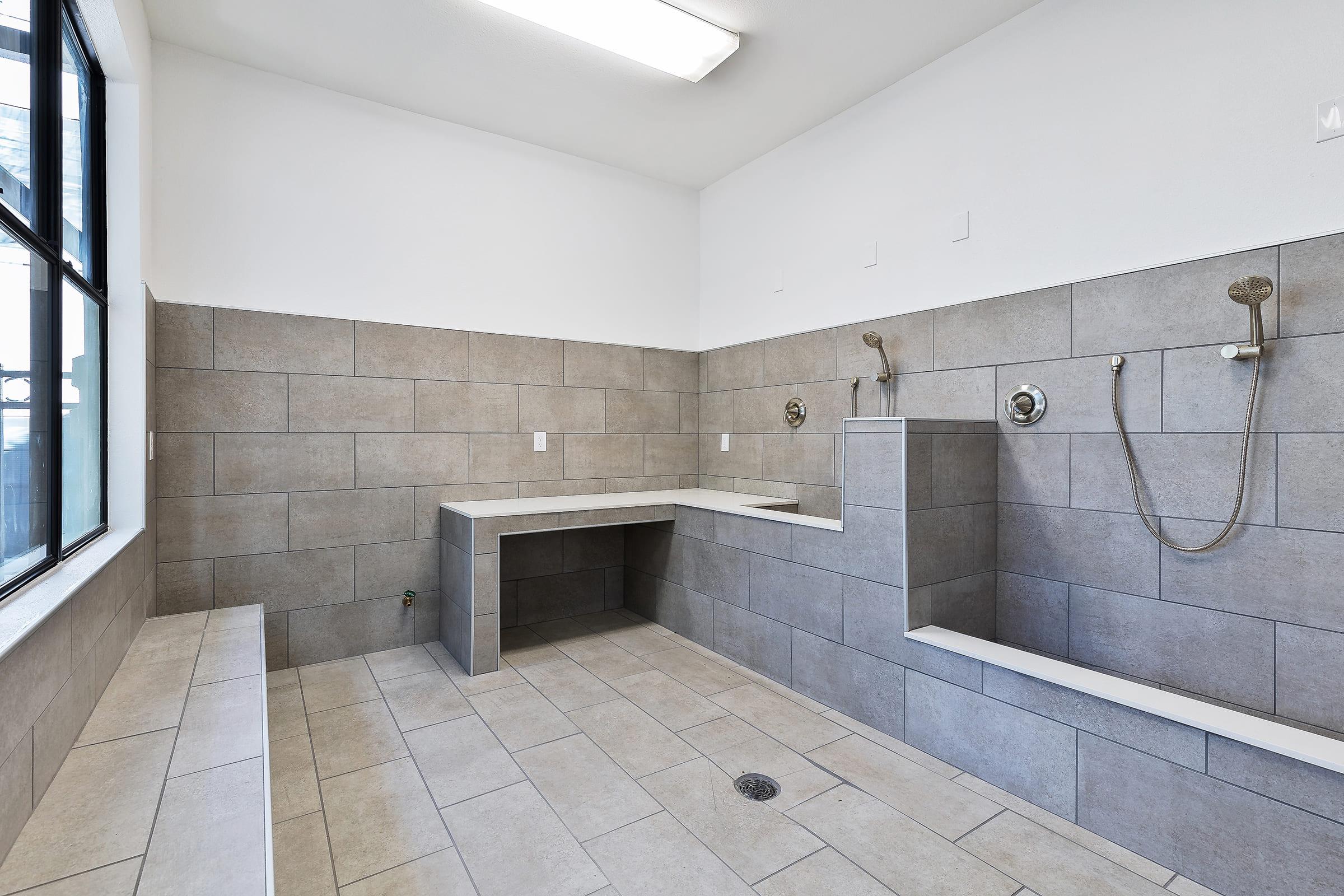 A modern, spacious bathroom with gray tiled walls and floors. Features include a shower area with two showerheads, a built-in bench, and a counter space along the wall. Natural light enters through a large window, providing a bright and clean atmosphere.