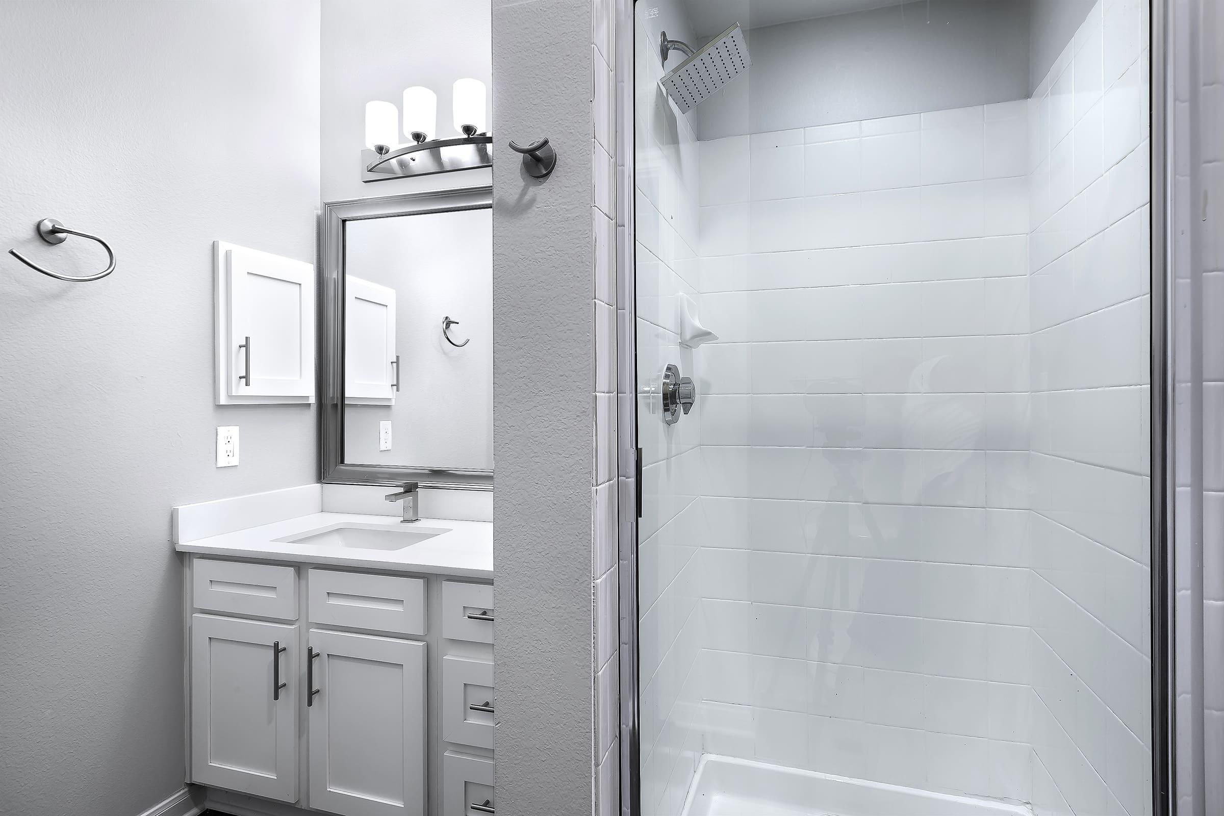 A modern bathroom featuring a glass-enclosed shower and a white vanity with a sink. The walls are painted in light grey, and there is a mirror above the vanity. Four light fixtures are mounted above the mirror. The shower area is clean and minimalist with white tiles.