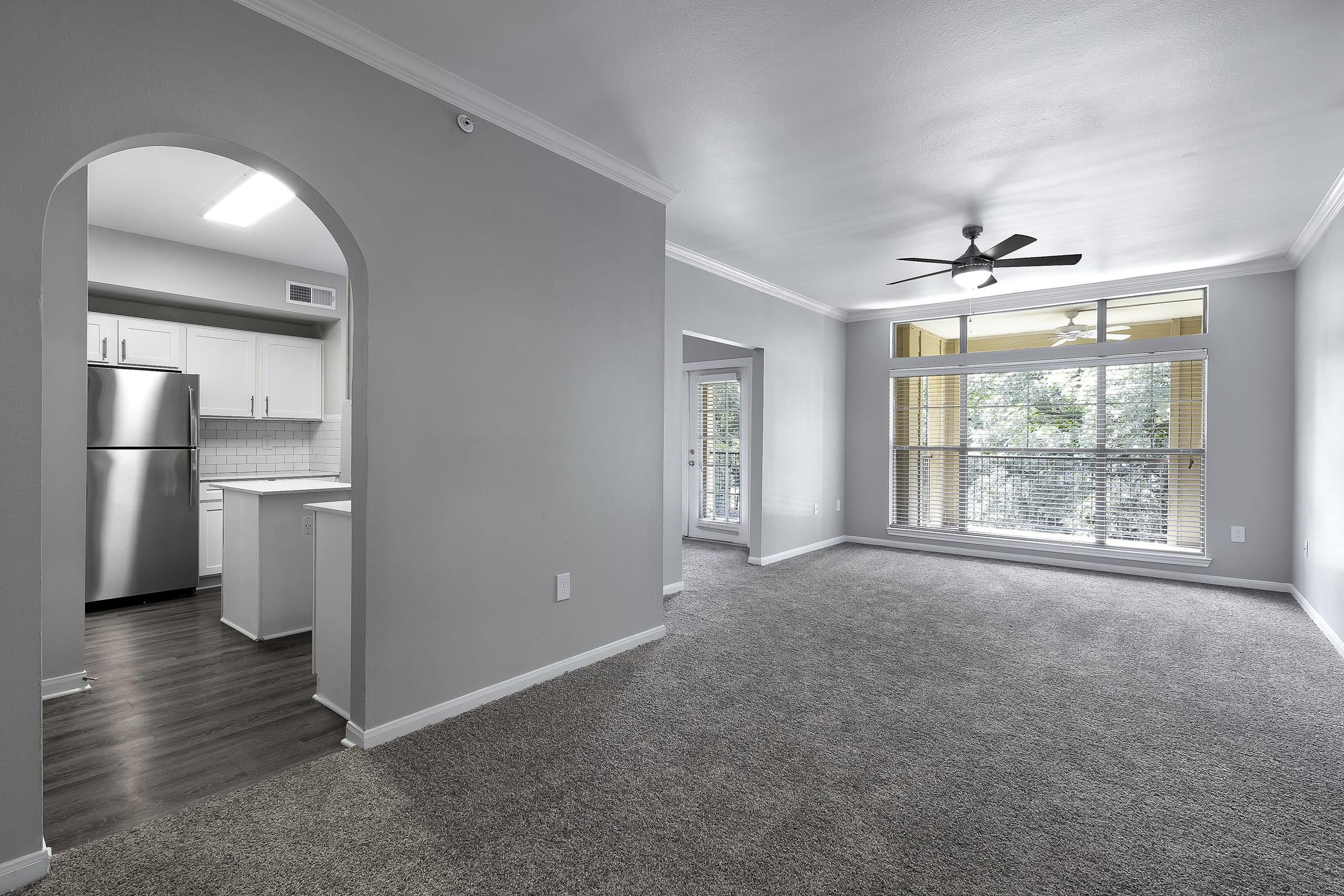 Spacious living area with light gray walls and carpet, featuring a large window with blinds. An archway leads to a modern kitchen with stainless steel appliances. Natural light fills the room, highlighting the open layout and ceiling fan for comfort.