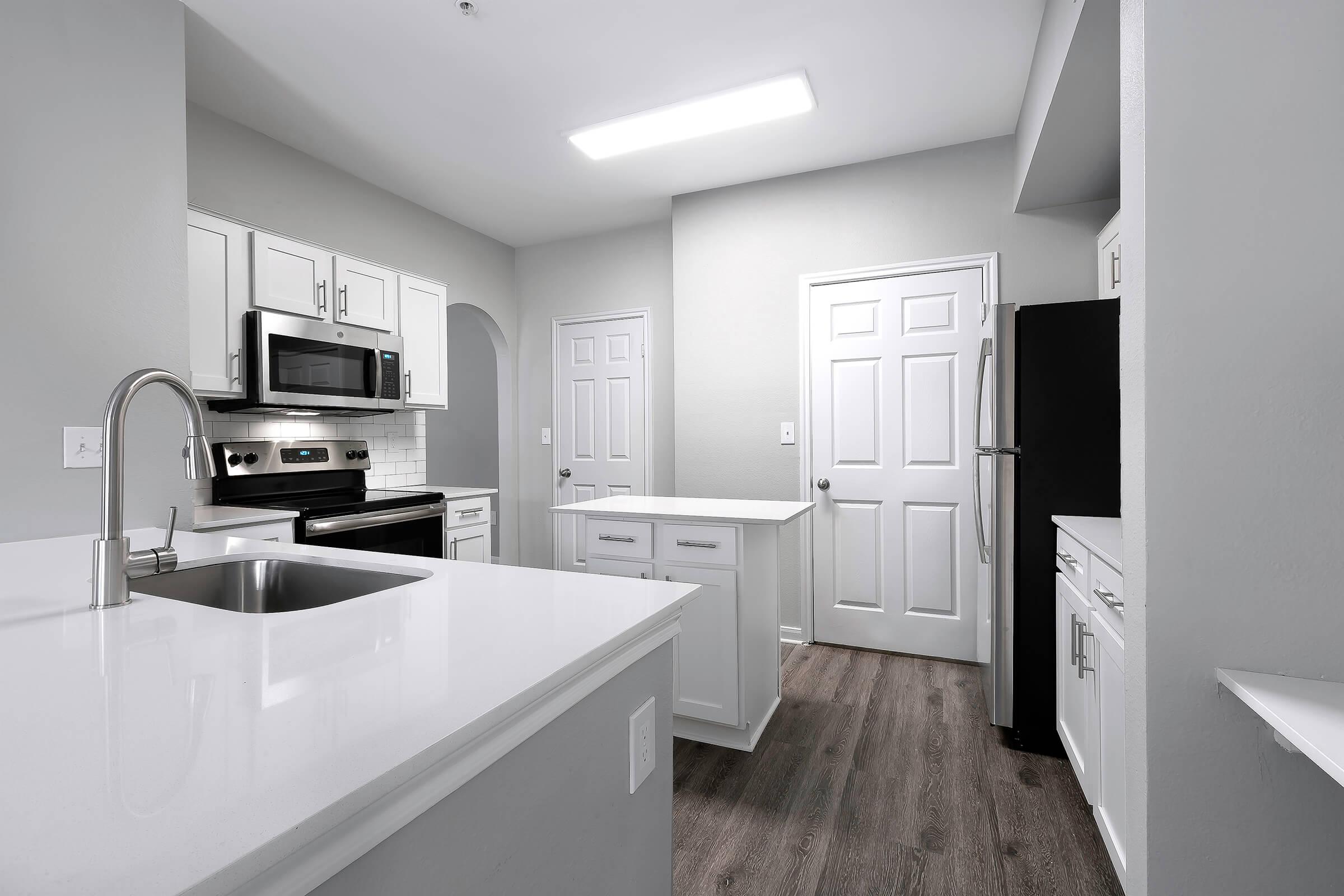 A modern kitchen featuring white cabinets, stainless steel appliances, and a large island with a sink. The walls are painted a light gray, and the floor is a wood-like finish. There are two doors leading to other rooms, and a bright overhead light illuminates the space.