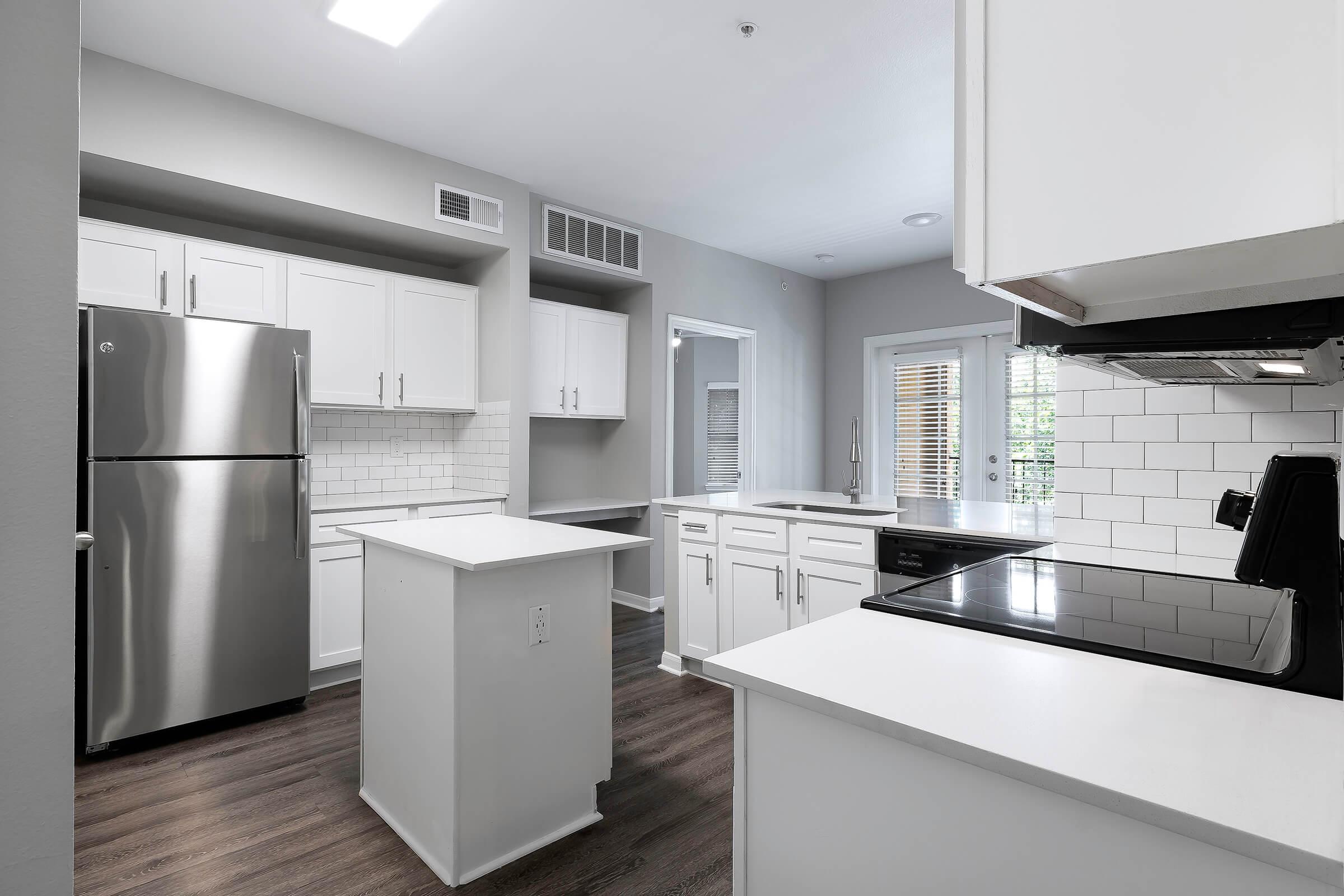 A modern kitchen featuring stainless steel appliances, including a refrigerator and an electric stove. The cabinetry is white, with a subway tile backsplash. An island is positioned in the center, and there are large windows providing natural light. The flooring is a dark wood laminate, enhancing the contemporary design.