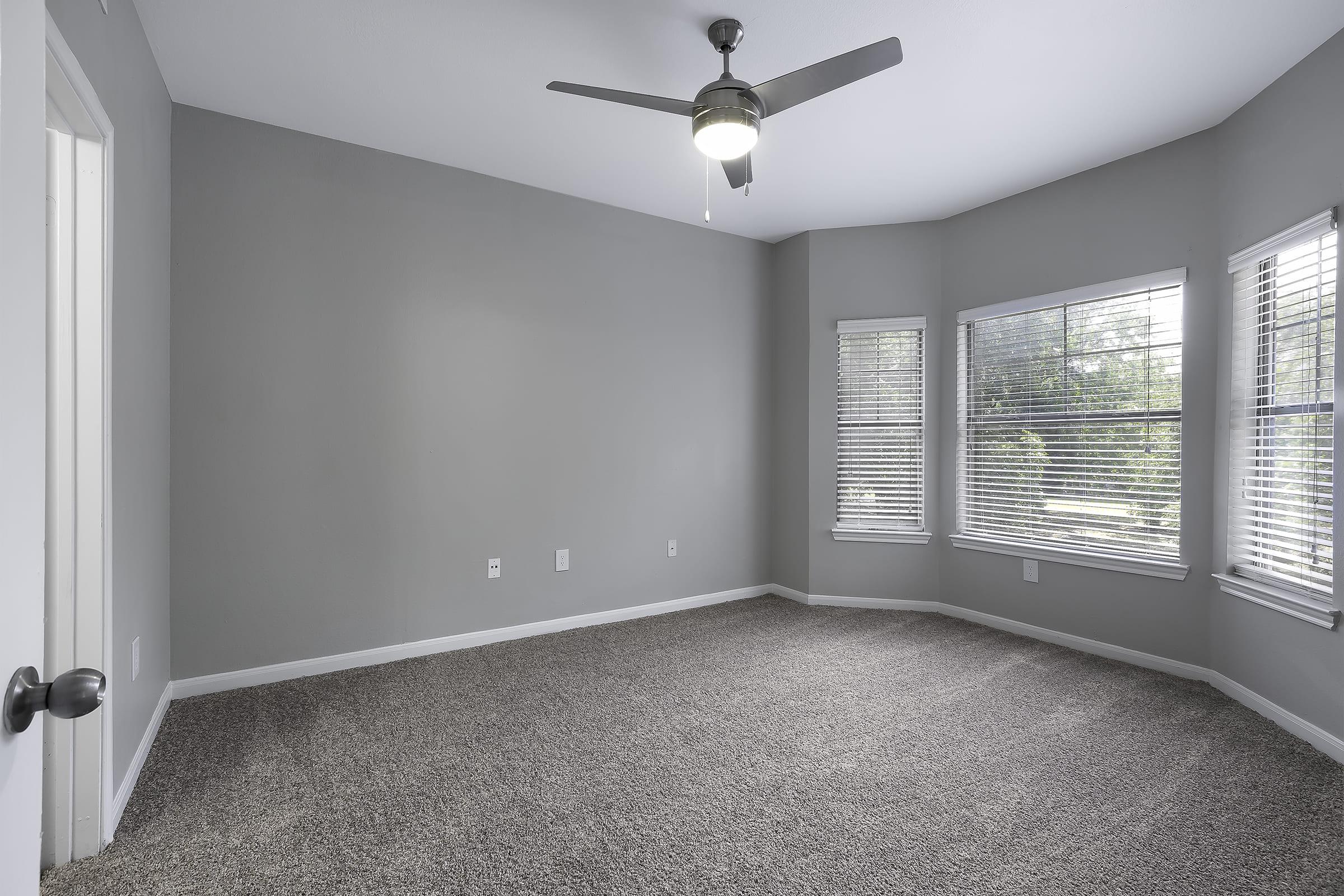 A spacious, empty room with gray walls and carpet flooring. It features a ceiling fan with a light fixture, and large windows with blinds allowing natural light to enter. The room is unadorned, providing a blank canvas for potential furnishings and decorations.