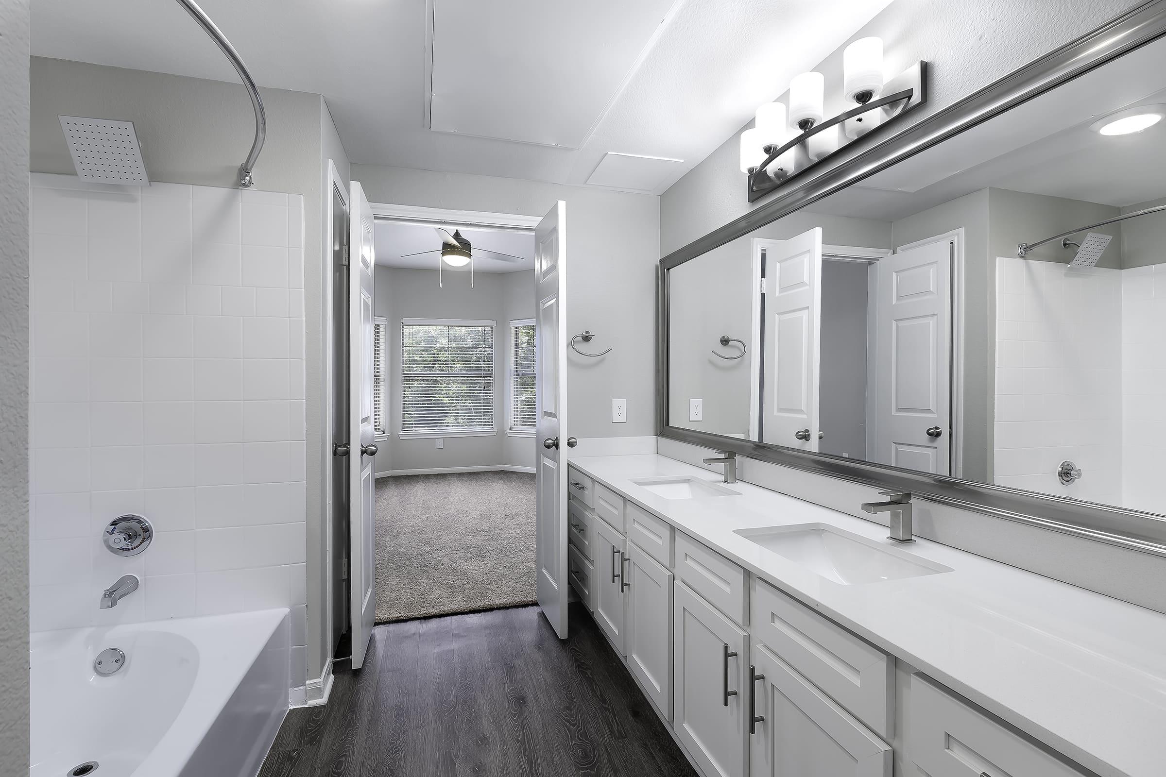 A modern bathroom featuring double sinks with a sleek countertop, a bathtub, and a shower area. The room has light gray walls, a large mirror above the sinks, and two open doors leading to a well-lit bedroom with carpeted flooring and windows.