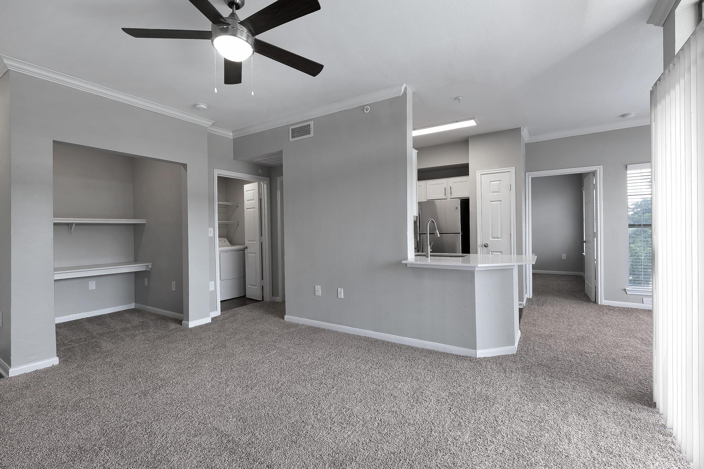 A spacious, modern apartment interior featuring light gray walls, carpeted flooring, and a ceiling fan. There are open areas including a kitchen with a bar counter, a washer/dryer nook, and a large window providing natural light. Doors lead to additional rooms, creating a bright and inviting atmosphere.