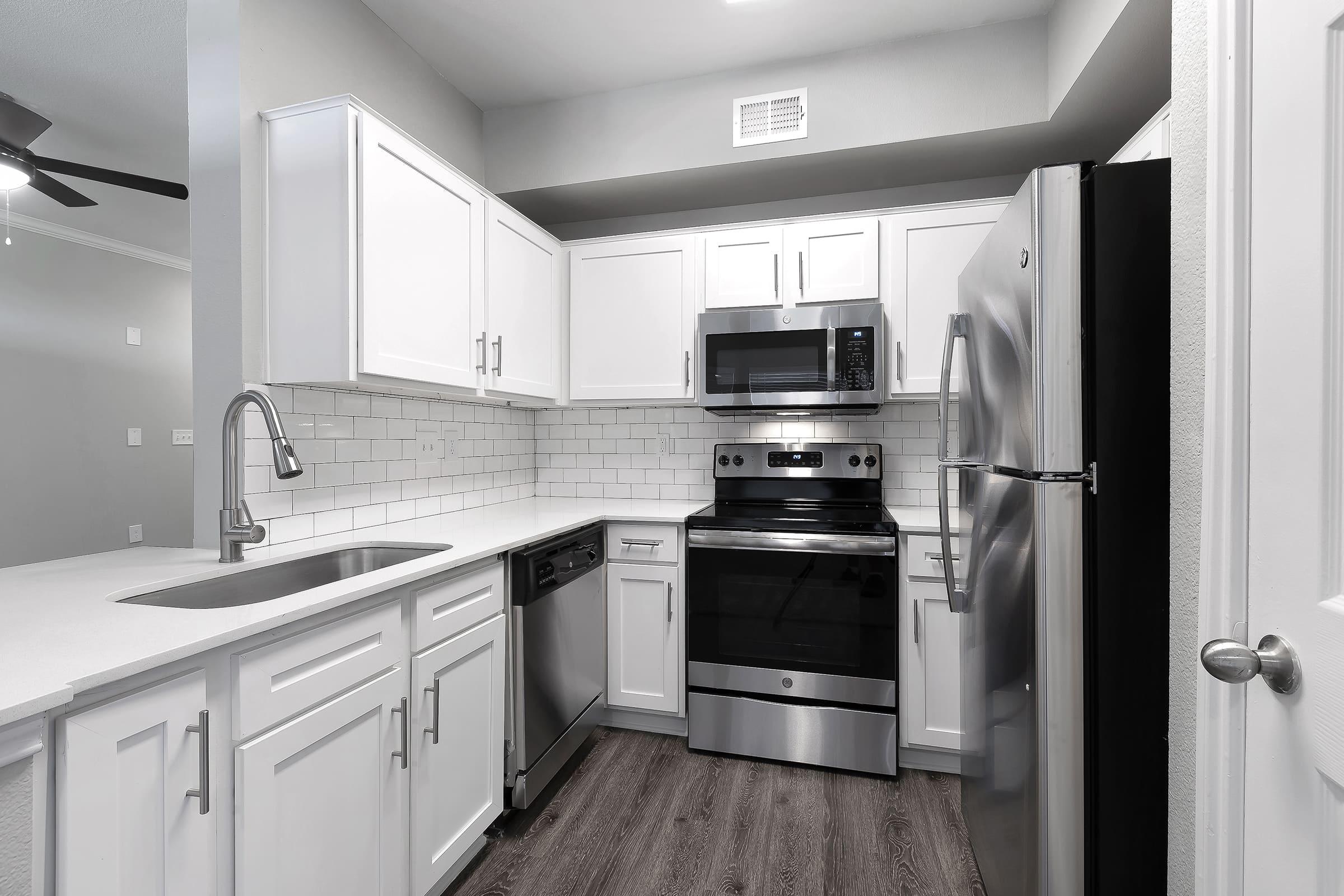 Modern kitchen featuring white cabinetry, stainless steel appliances including a refrigerator, microwave, range, and dishwasher. The countertop is smooth and white, complemented by a backsplash of white tiles. Wood-like flooring enhances the contemporary design, and a ceiling fan is visible.