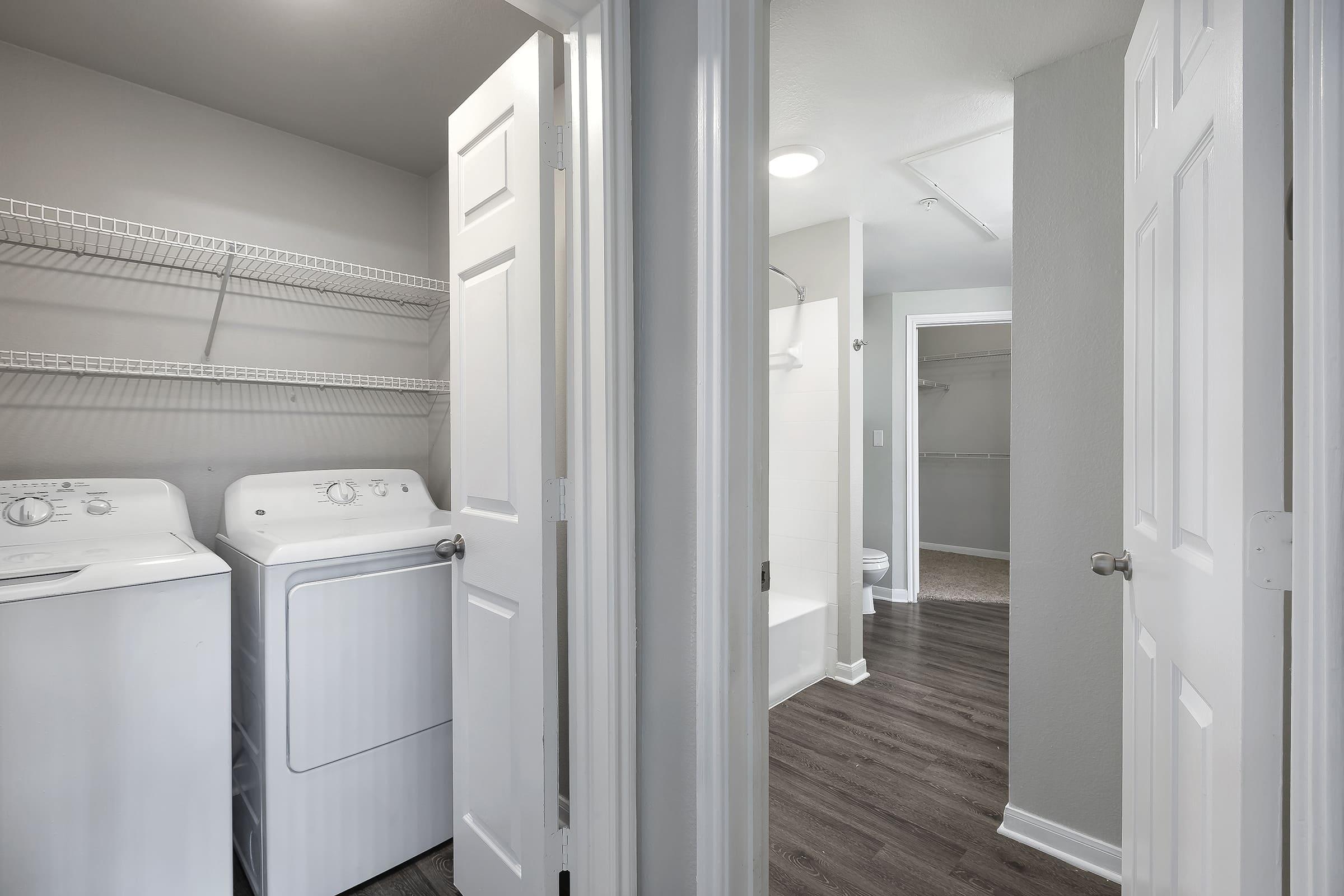 A bright laundry room featuring a washer and dryer side by side, with white shelving above. Two open doors lead to other areas—one entering a bathroom with a shower and the other into a closet space. The floor is covered in light brown laminate, and the walls are painted a soft gray.