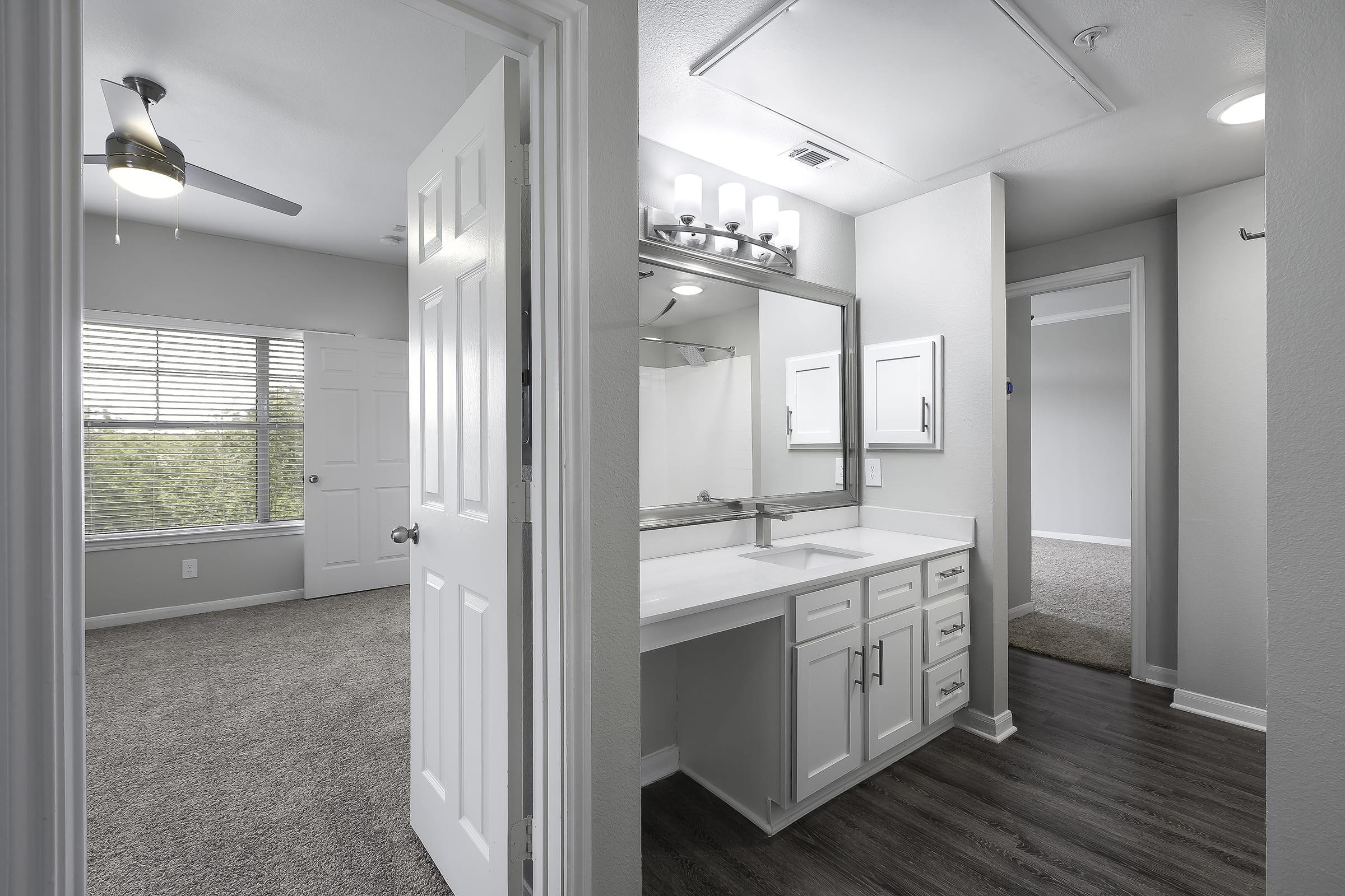 Interior view of a modern apartment showing a bathroom vanity with a large mirror and light fixtures. In the background, there is a door leading to a bedroom with a window, allowing natural light, and a ceiling fan. The flooring is a combination of carpet and laminate.