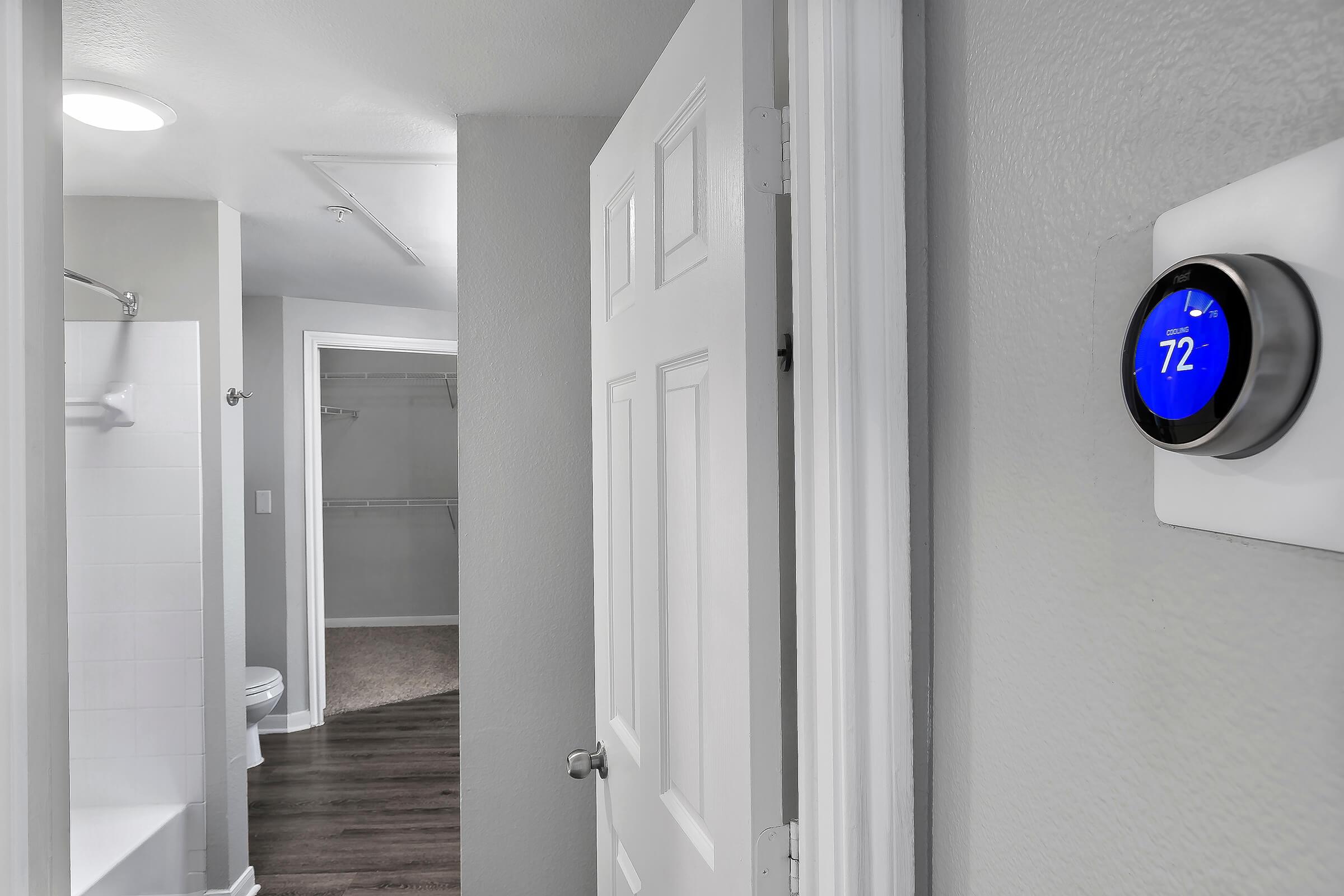 Interior view of a bathroom area. A white door leads to the bathroom with a shower and closet visible. A modern thermostat is mounted on the wall, displaying a temperature of 72°F. The walls are painted light gray, and the floor features dark wood laminate, creating a clean and contemporary atmosphere.